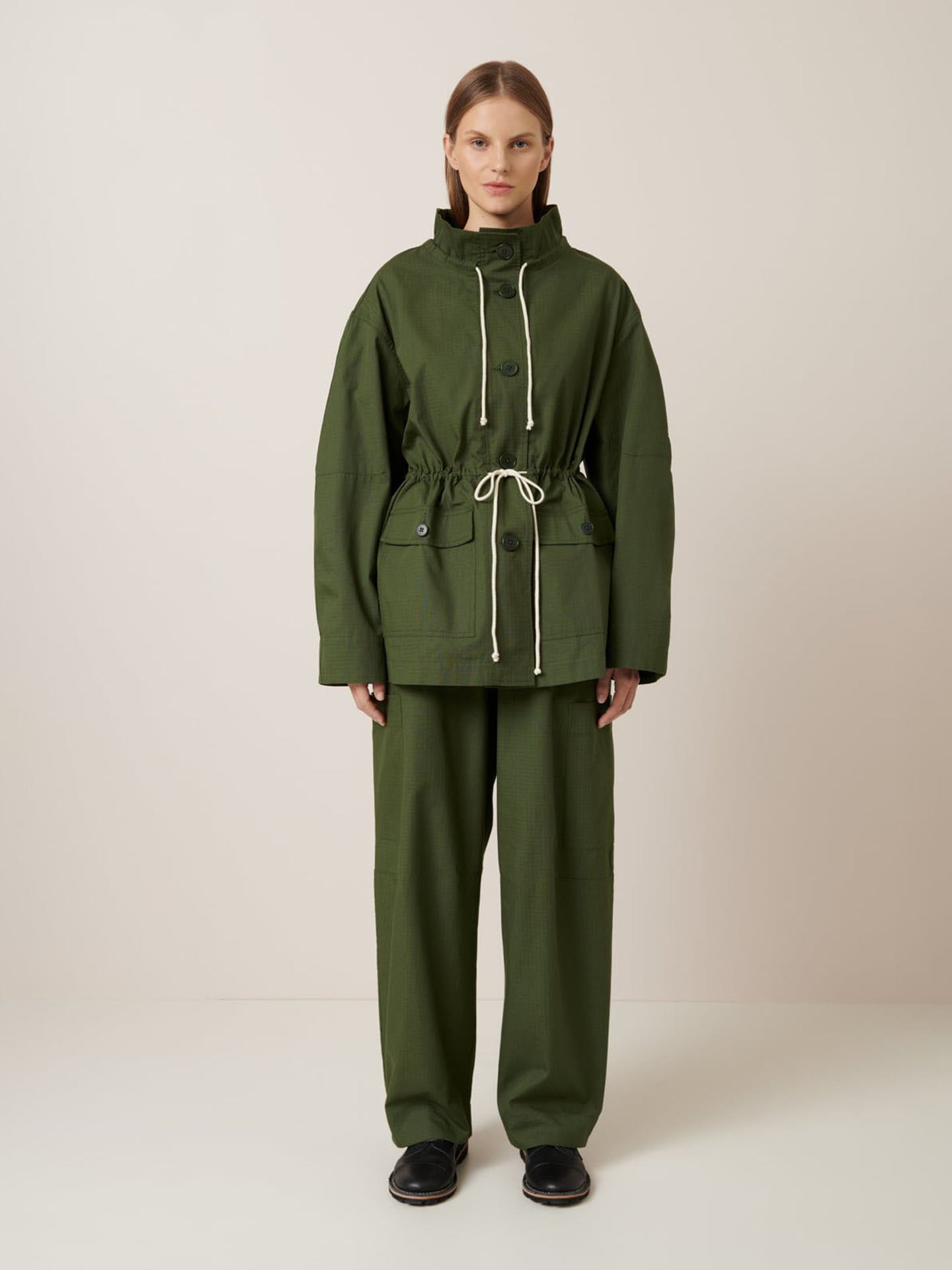 A woman faces forward in the Kowtow Echo Jacket – Forest, an oversized olive green jacket with drawstrings and matching pants, paired with black shoes. The background is plain and neutral.