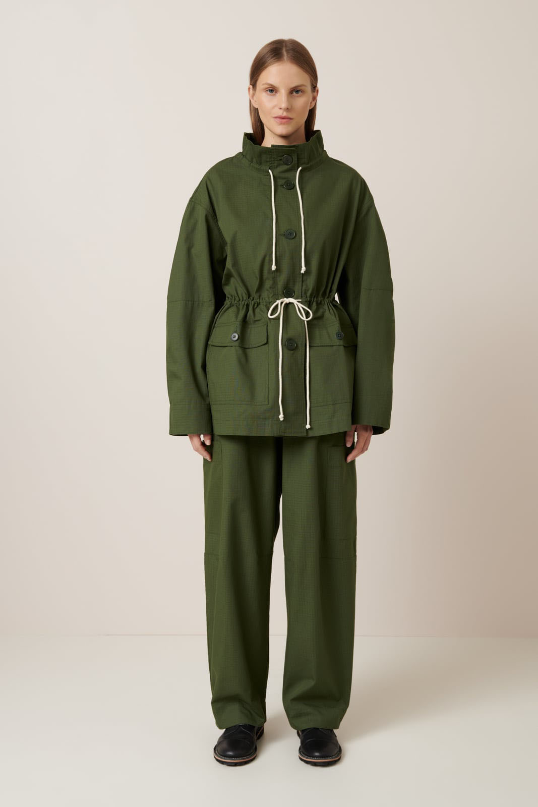 A woman faces forward in the Kowtow Echo Jacket – Forest, an oversized olive green jacket with drawstrings and matching pants, paired with black shoes. The background is plain and neutral.