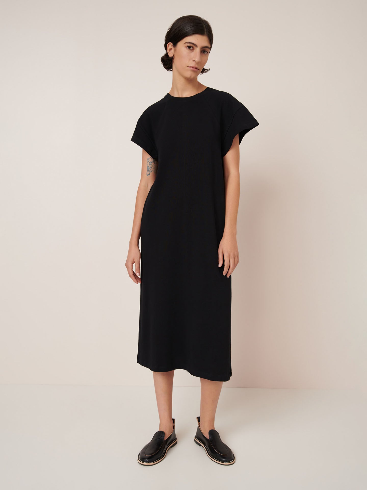A person stands against a plain background wearing the Kowtow Effect Dress – Black, a loose-fitting, standard fit midi dress, paired with black slip-on shoes.