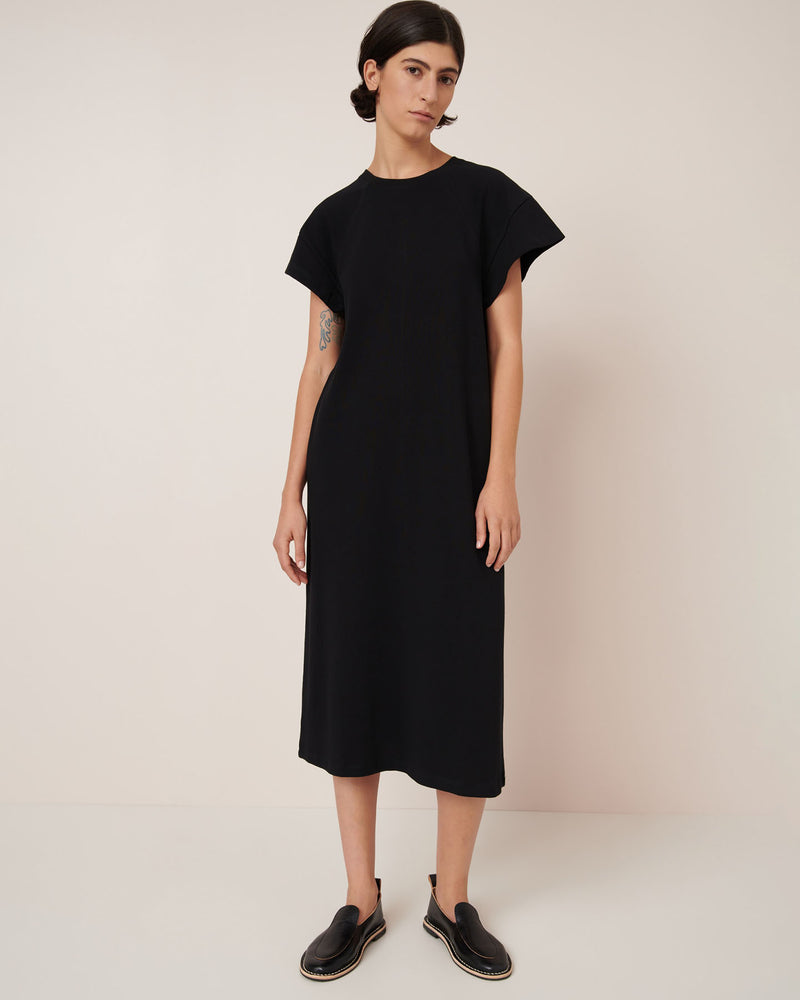 A person stands against a plain background wearing the Kowtow Effect Dress – Black, a loose-fitting, standard fit midi dress, paired with black slip-on shoes.