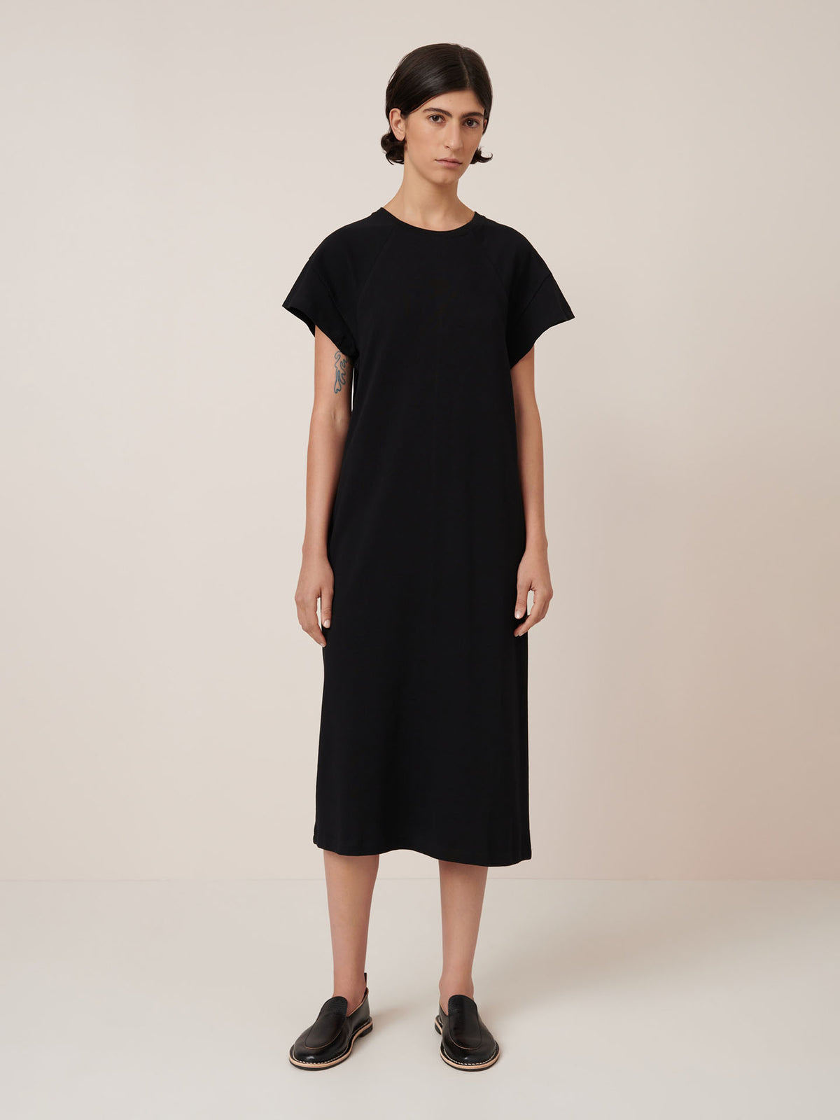 A person stands against a plain background, wearing the Effect Dress – Black by Kowtow, a loose-fitting midi dress paired with black slip-on shoes that showcase the brand’s signature standard fit.