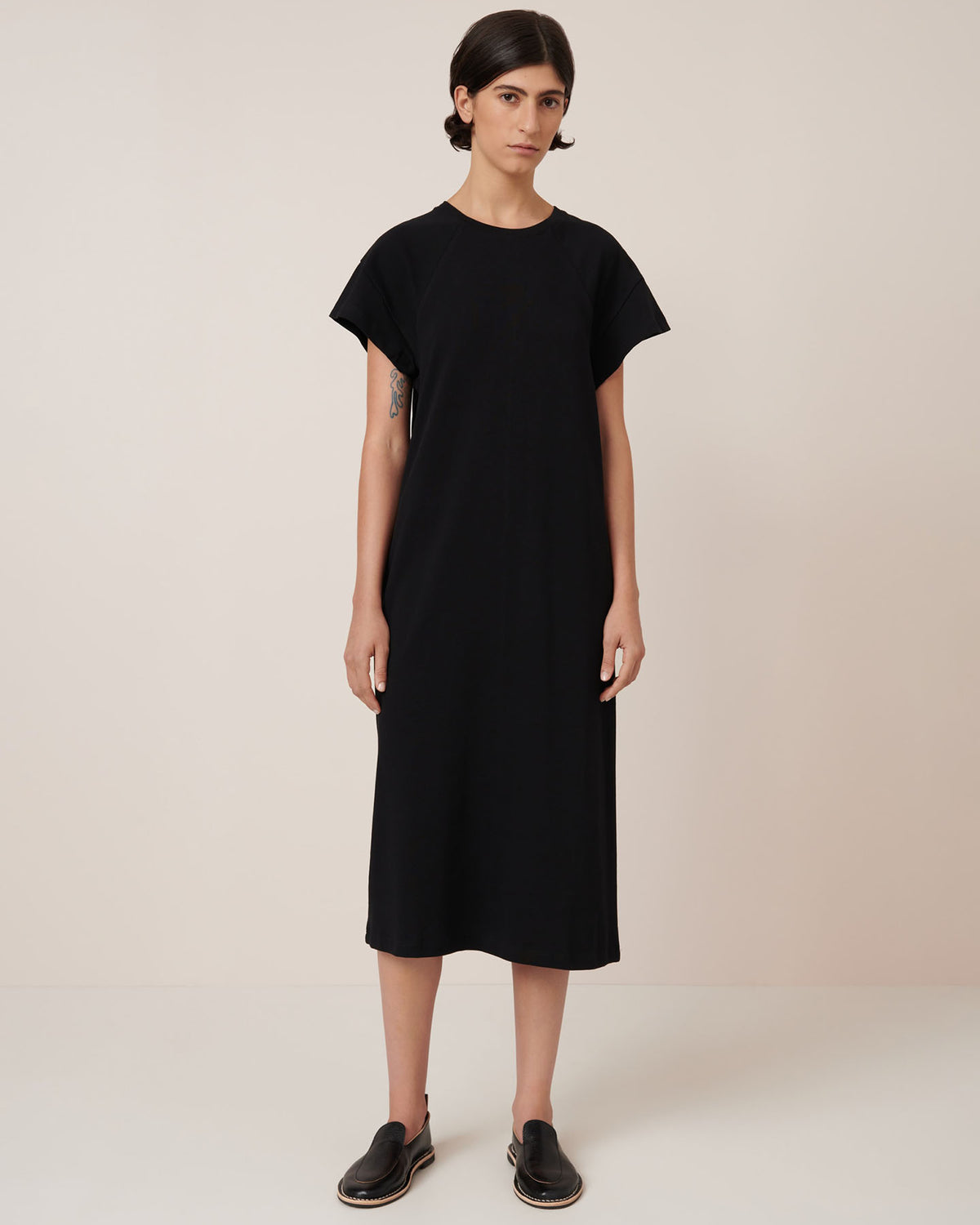 A person stands against a plain background, wearing the Effect Dress – Black by Kowtow, a loose-fitting midi dress paired with black slip-on shoes that showcase the brand’s signature standard fit.