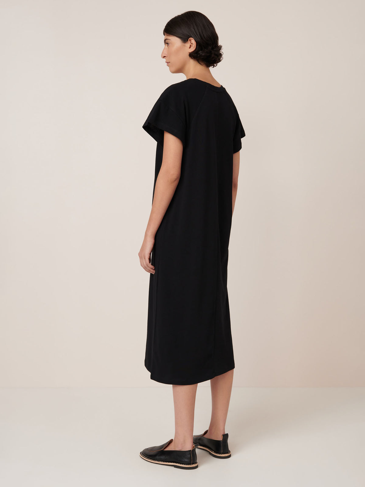 A person stands sideways against a plain background wearing the Kowtow Effect Dress – Black, a loose-fitting, short-sleeve midi dress styled with black slip-on shoes, highlighting its relaxed standard fit.