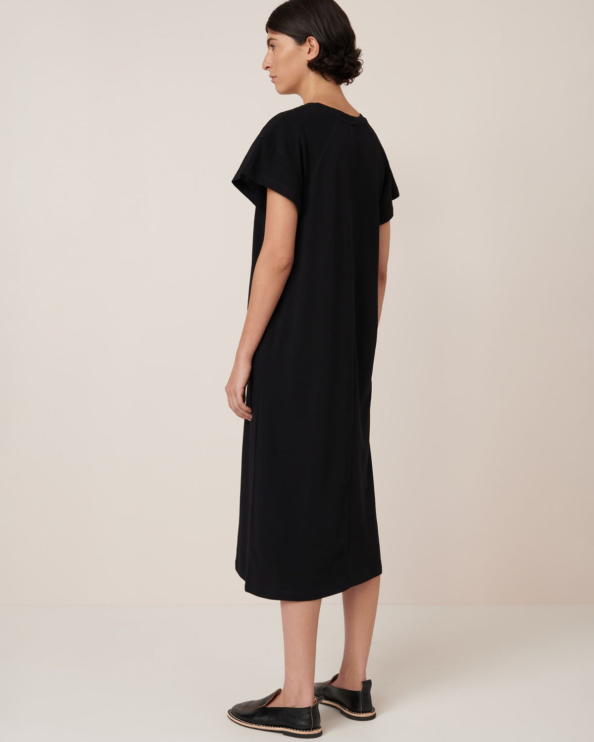 A person stands sideways against a plain background wearing the Kowtow Effect Dress – Black, a loose-fitting, short-sleeve midi dress styled with black slip-on shoes, highlighting its relaxed standard fit.