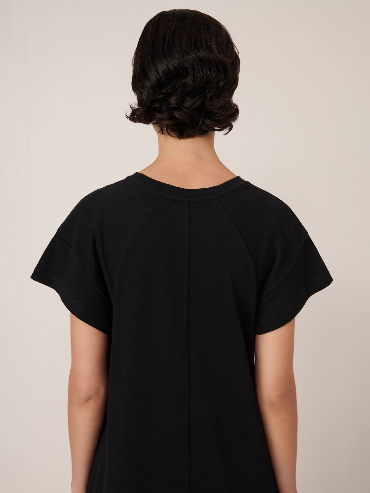 A person with short, dark, wavy hair is shown from the back against a neutral background wearing the Effect Dress in Black by Kowtow, showcasing the brand&#39;s standard fit and precise garment measurements.