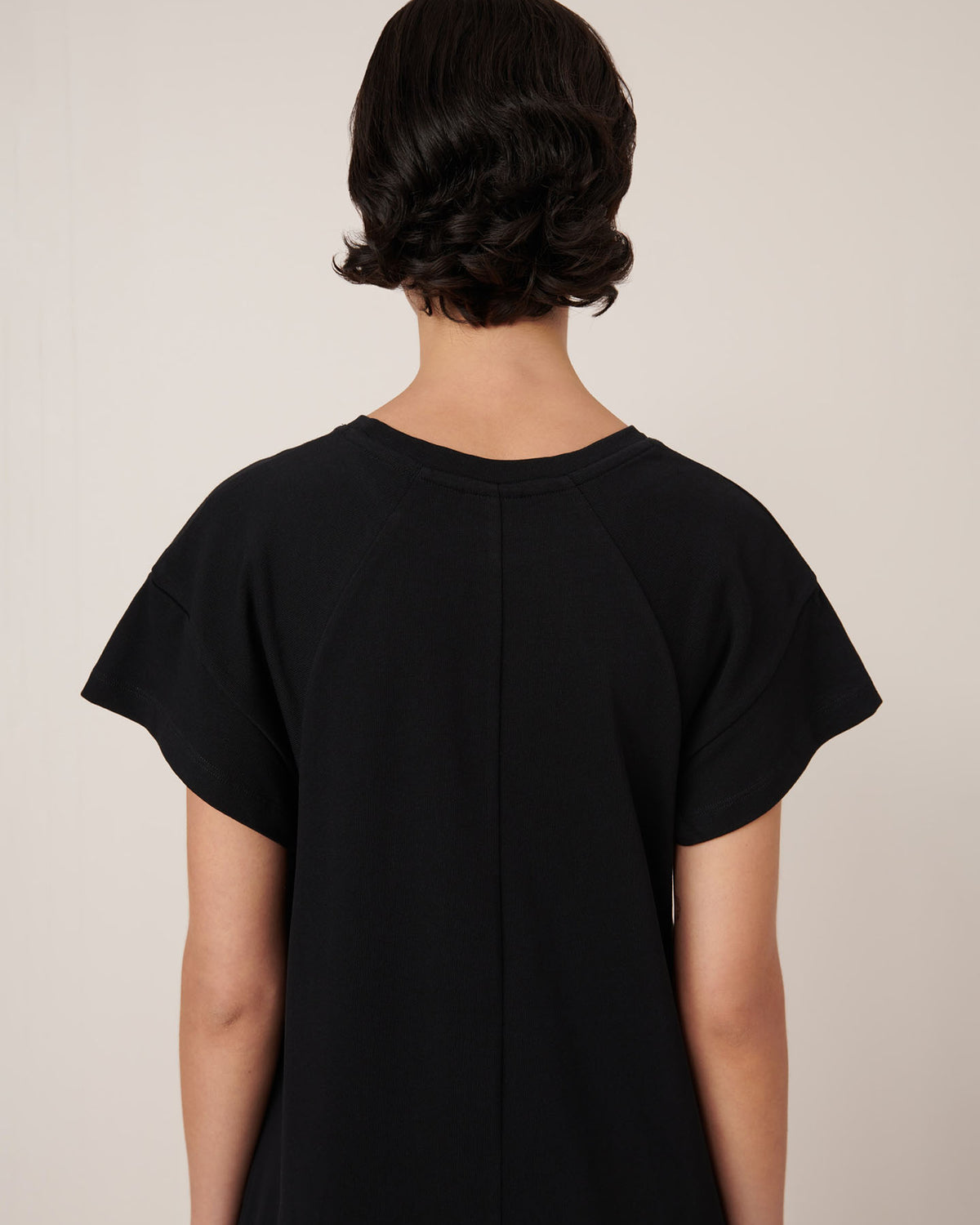 A person with short, dark, wavy hair is shown from the back against a neutral background wearing the Effect Dress in Black by Kowtow, showcasing the brand's standard fit and precise garment measurements.