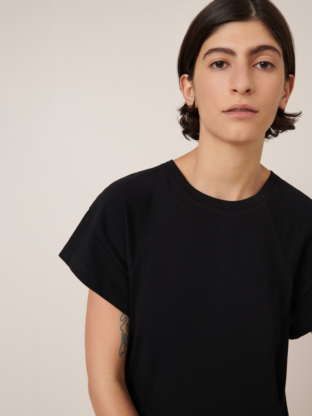 A person with short dark hair wearing the Kowtow Effect Dress in black, standing against a neutral light background, with a tattoo partially visible on their left arm.