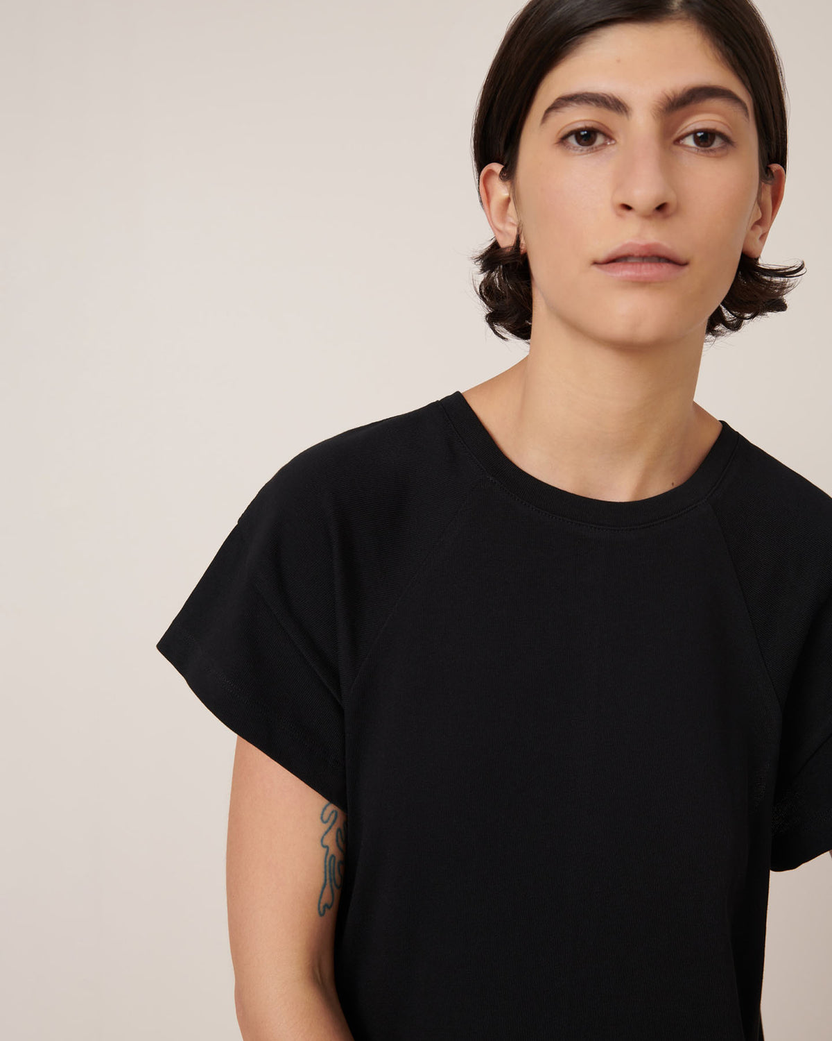 A person with short dark hair wearing the Kowtow Effect Dress in black, standing against a neutral light background, with a tattoo partially visible on their left arm.