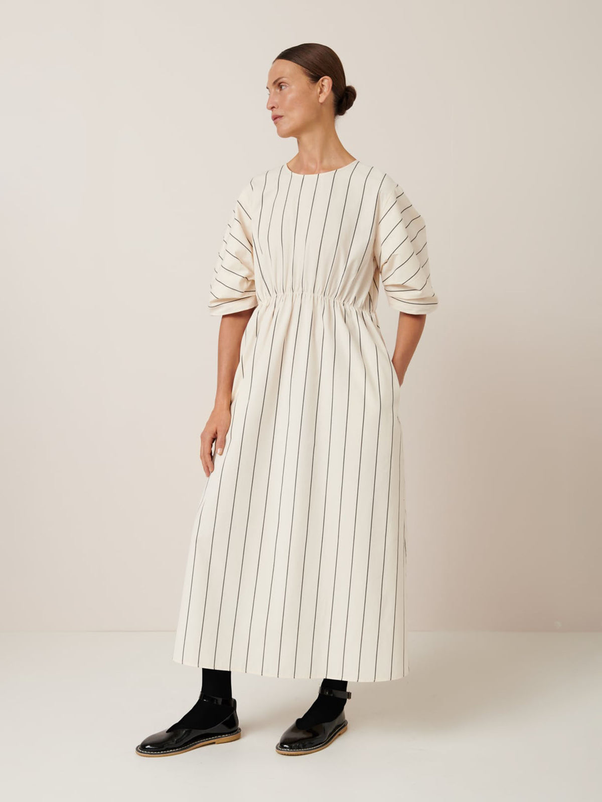 In the Ellora Dress – Greige Pinstripe by Kowtow, a person stands against a plain background. The long, relaxed fit dress features vertical stripes and short puff sleeves. Paired with black shoes and socks, they gaze aside, embodying effortless simplicity through Kowtow&#39;s design.