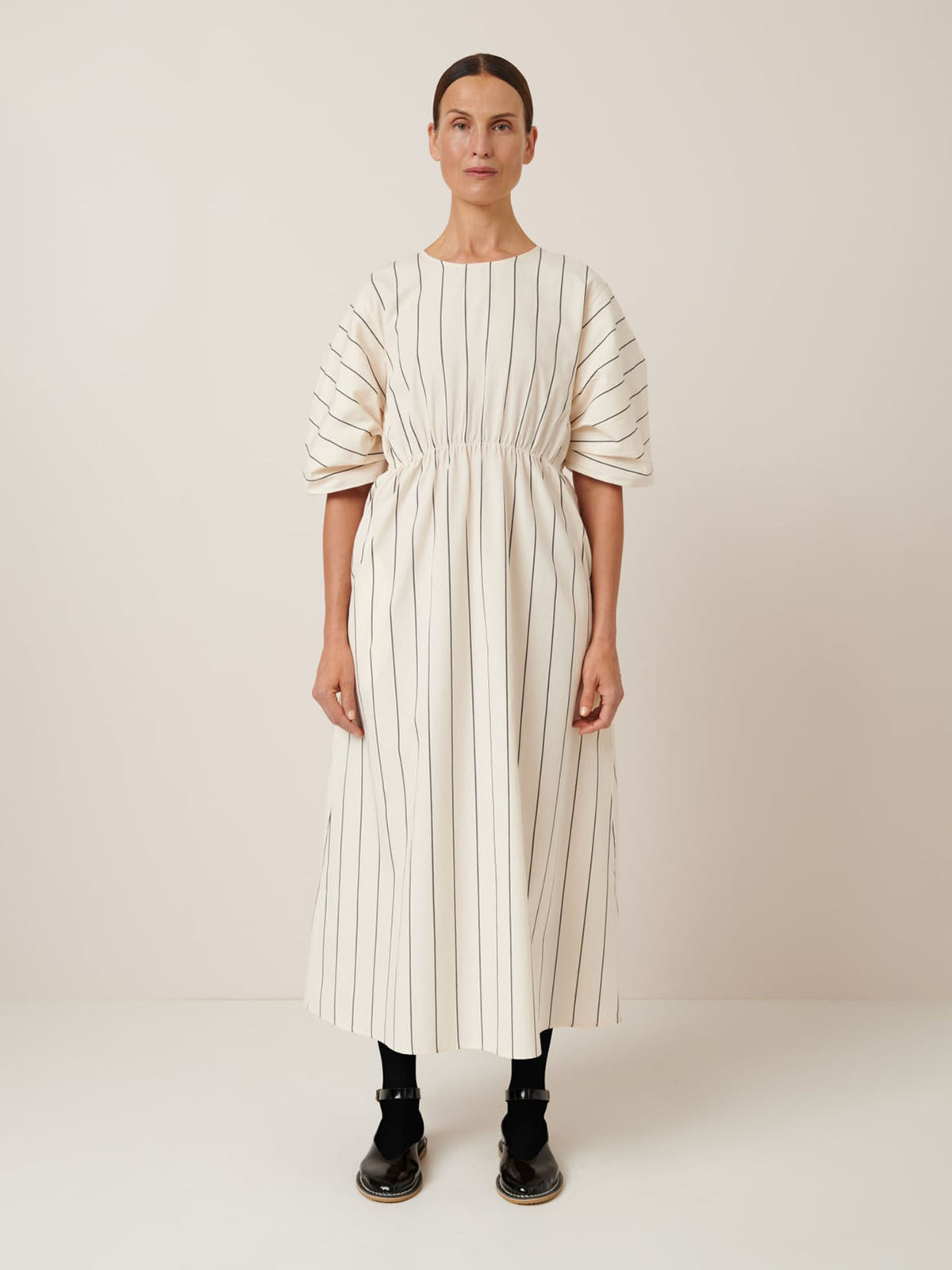 A person stands against a beige background in the Kowtow Ellora Dress – Greige Pinstripe, featuring black vertical stripes and puffed sleeves for a relaxed fit. Styled with black sandals and socks, it exudes effortless style. Refer to our size guide for perfect measurements.