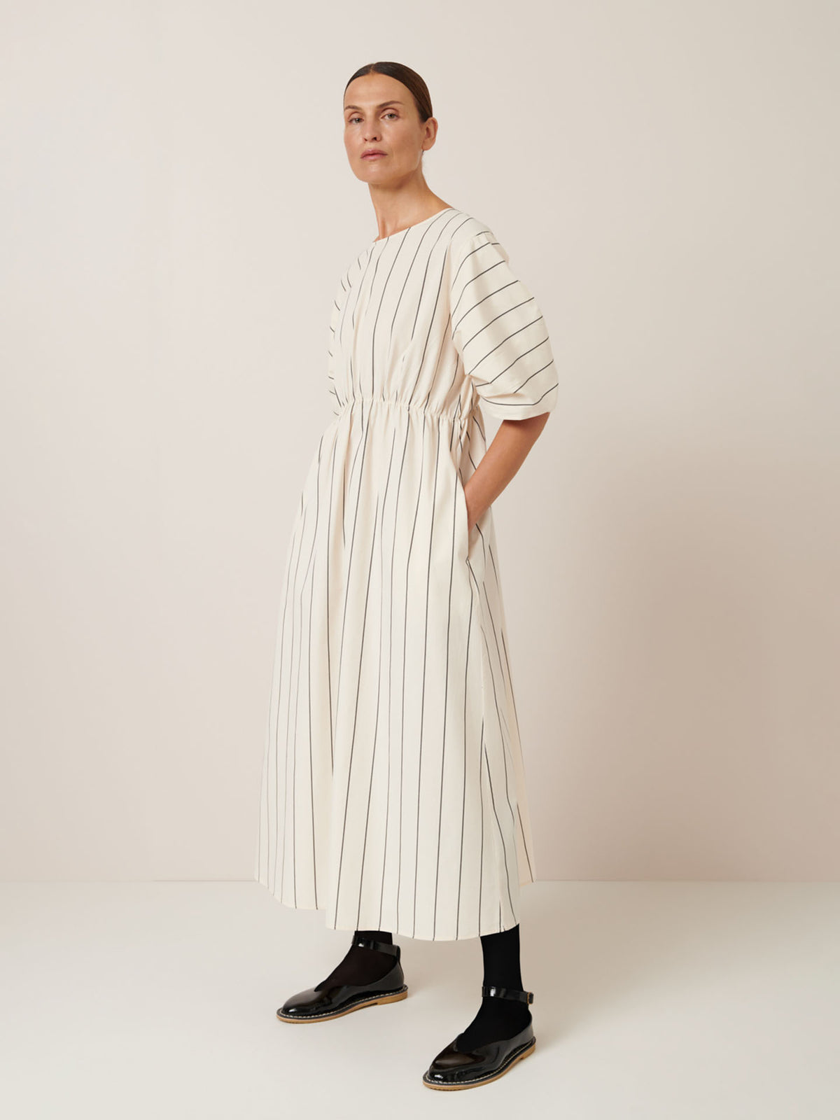 A person wears the Ellora Dress – Greige Pinstripe by Kowtow, set against a plain backdrop. Its relaxed fit pairs seamlessly with black socks and sandals, combining comfort and effortless style.