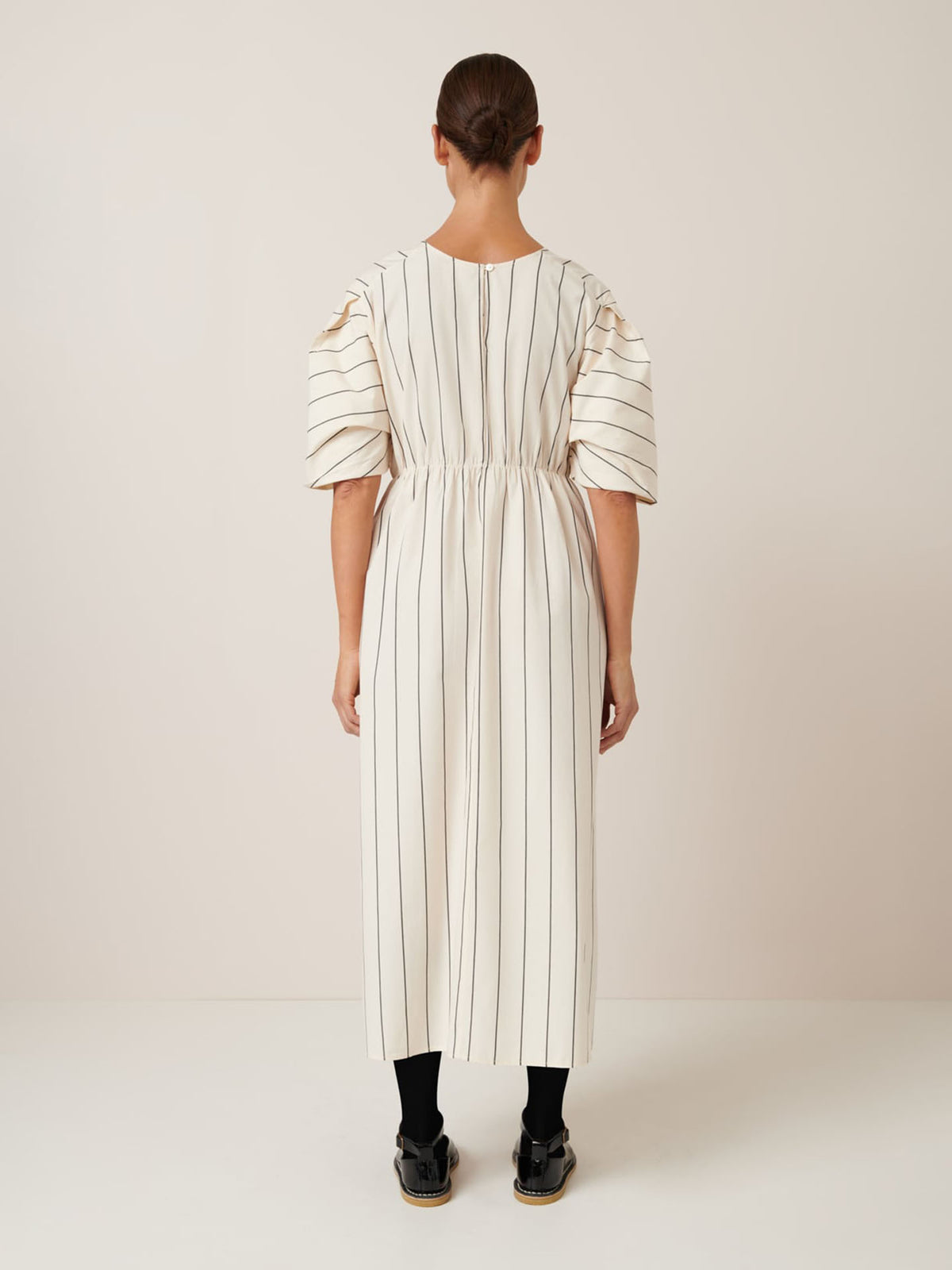 A person models the Ellora Dress by Kowtow, featuring a relaxed fit with black vertical stripes on greige and puffed sleeves. The back view against a plain backdrop showcases its flowing design.