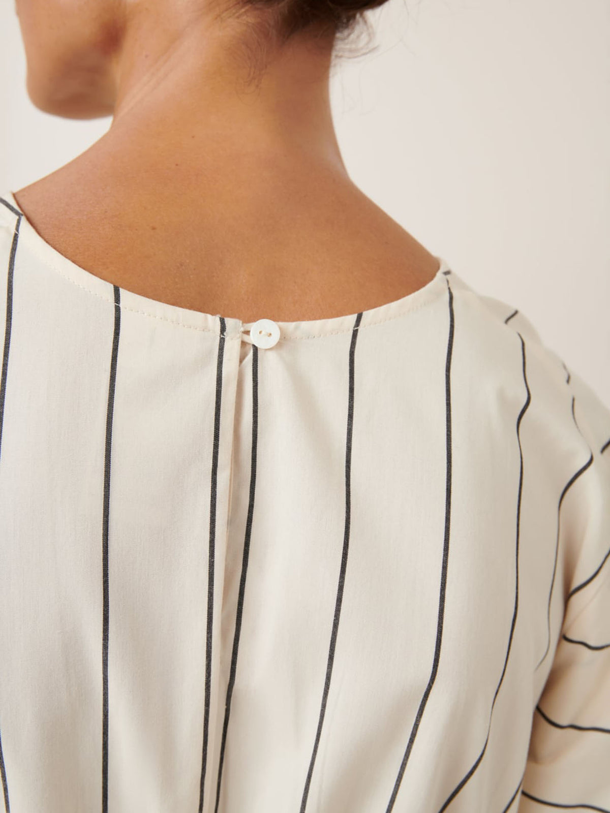 Close-up of a person in the Ellora Dress – Greige Pinstripe by Kowtow, highlighting its relaxed fit. A small button near the neckline adds elegance, as captured in our size guide for precise measurements.