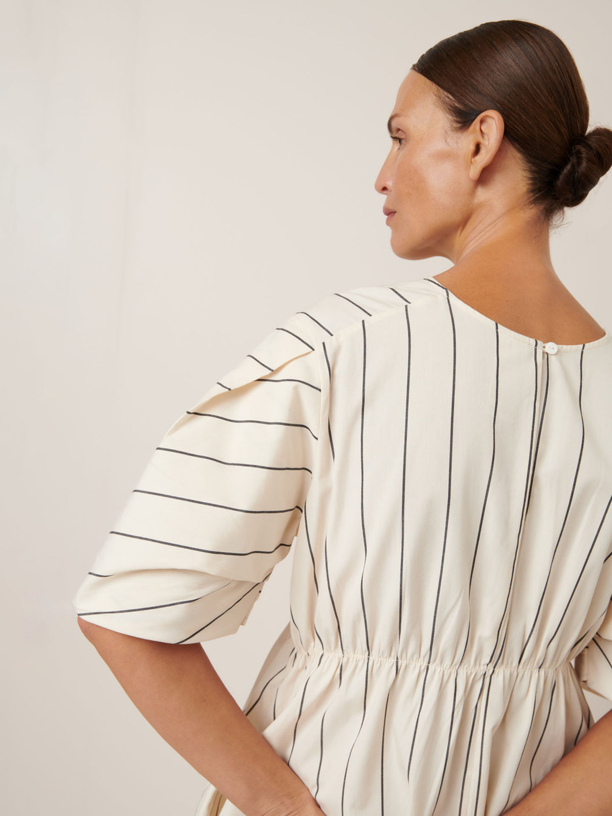 A person wearing the Kowtow Ellora Dress in Greige Pinstripe is seen from behind, with hair elegantly styled in a bun.