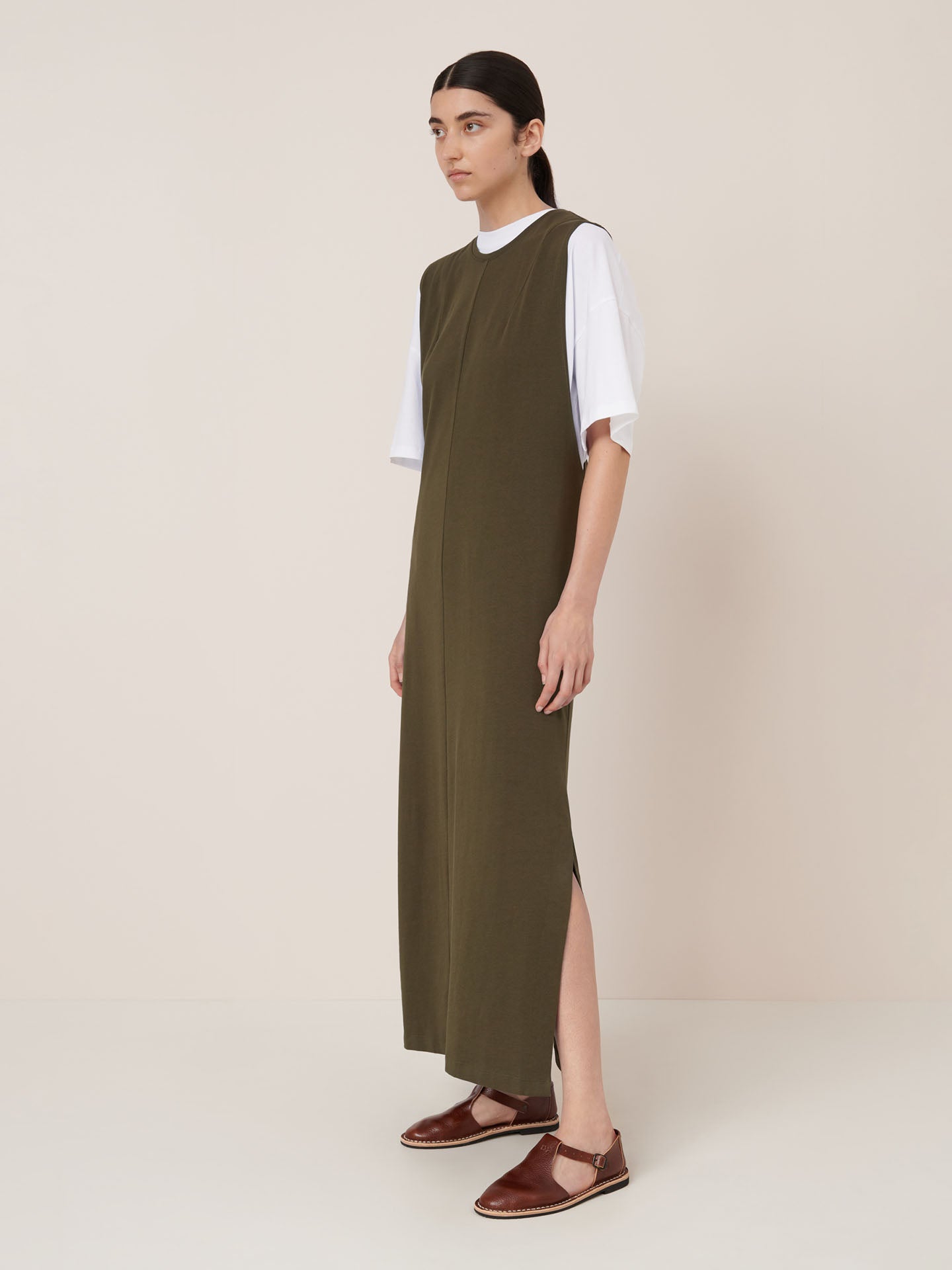A person stands against a plain background wearing the Kowtow Elm Dress – Ivy, a relaxed fit olive green maxi dress with side slits, layered over a white T-shirt and paired with brown flat shoes.
