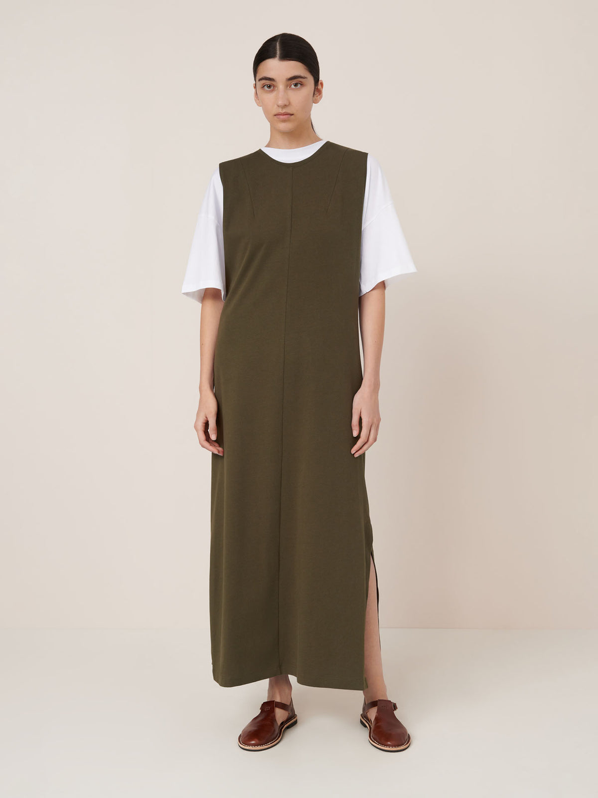 A person stands against a plain background wearing a white T-shirt under the Kowtow Elm Dress – Ivy, a relaxed-fit, olive green maxi dress with side slits, paired with brown sandals.