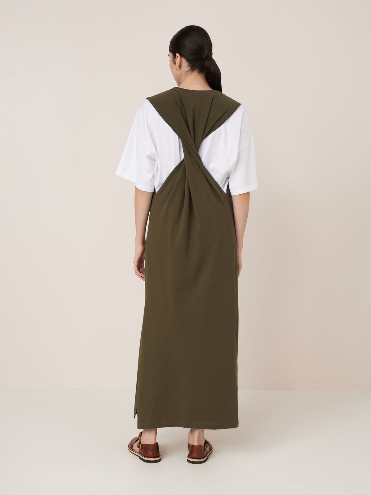 A person stands facing away, wearing the Kowtow Elm Dress in Ivy—an olive green, relaxed-fit, sleeveless dress with crisscrossed back—layered over a white t-shirt and paired with brown sandals.