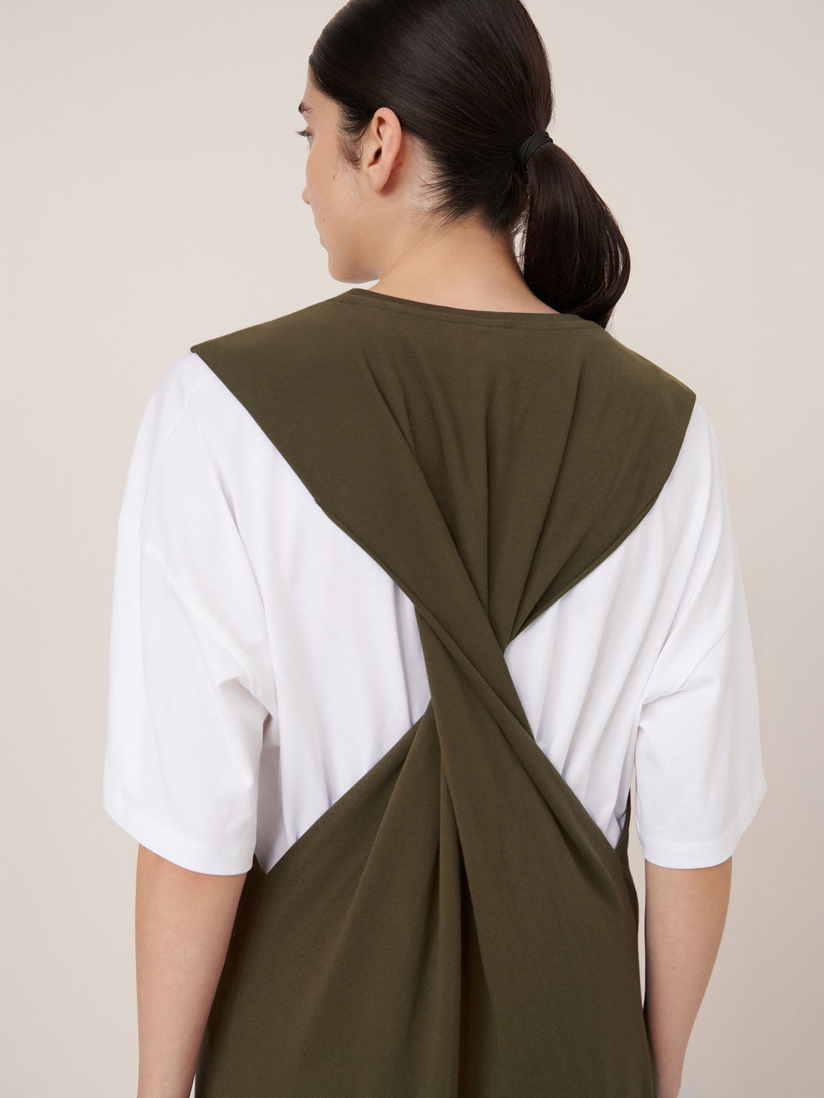 A person with long dark hair in a low ponytail wears the Kowtow Elm Dress – Ivy, featuring a white shirt base and olive green fabric twisted across the back for a signature relaxed fit.