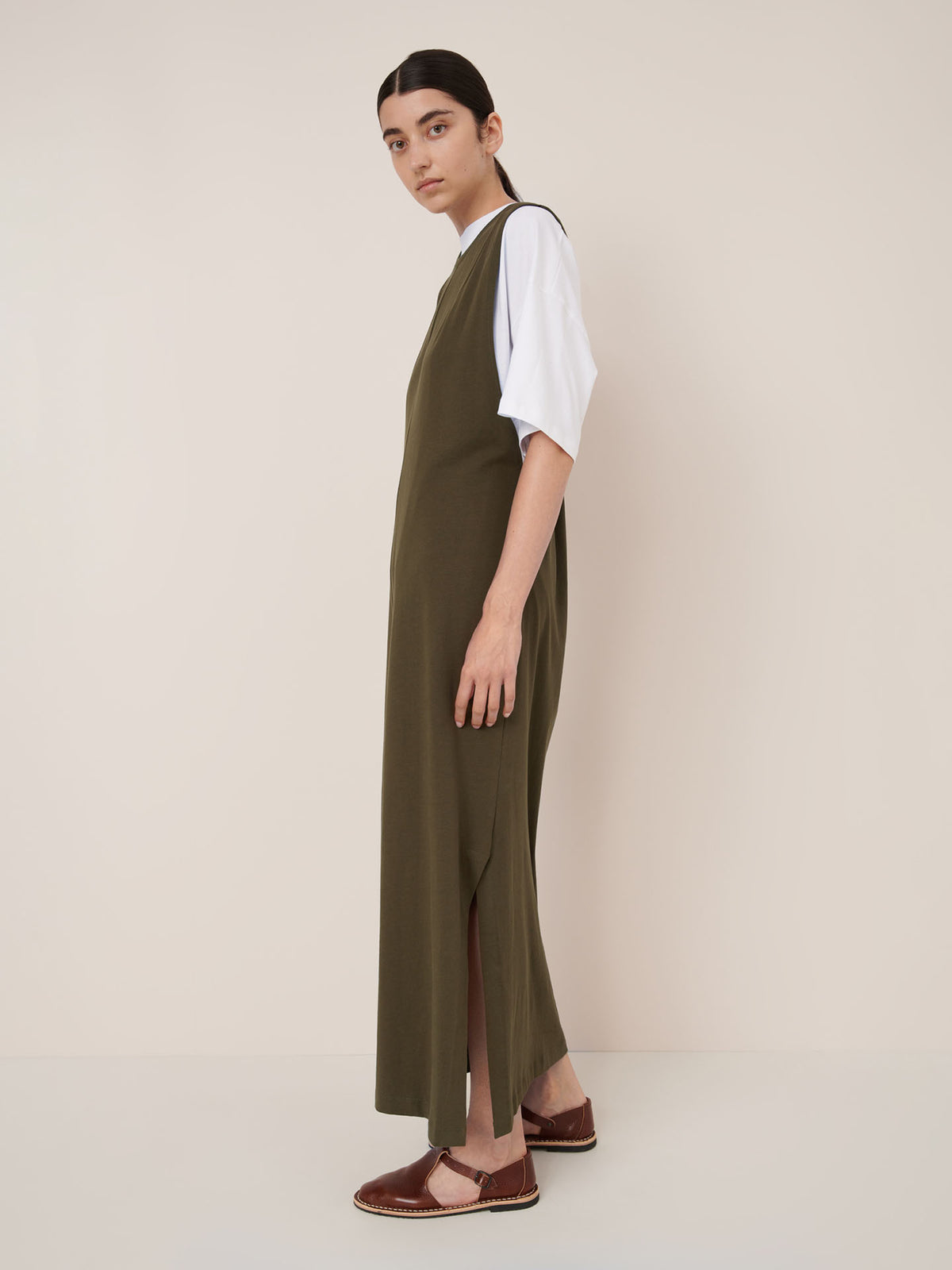 A person stands sideways in a relaxed fit Ivy green Elm Dress by Kowtow, layered over a white short-sleeve top and paired with brown flat shoes against a plain background.