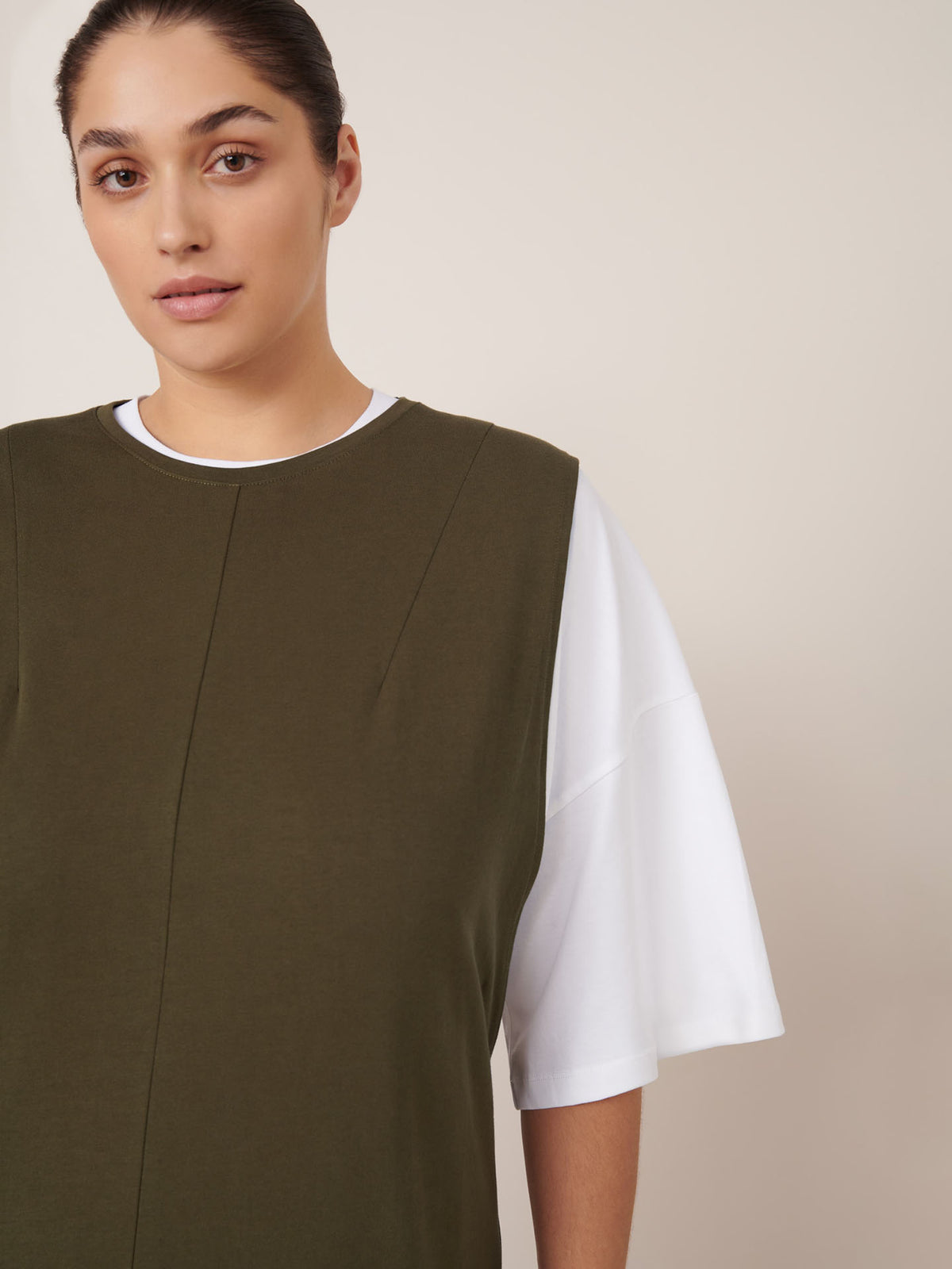 A person with dark hair pulled back is wearing the Kowtow Elm Dress in Ivy, styled in a relaxed fit, and stands against a plain light background.