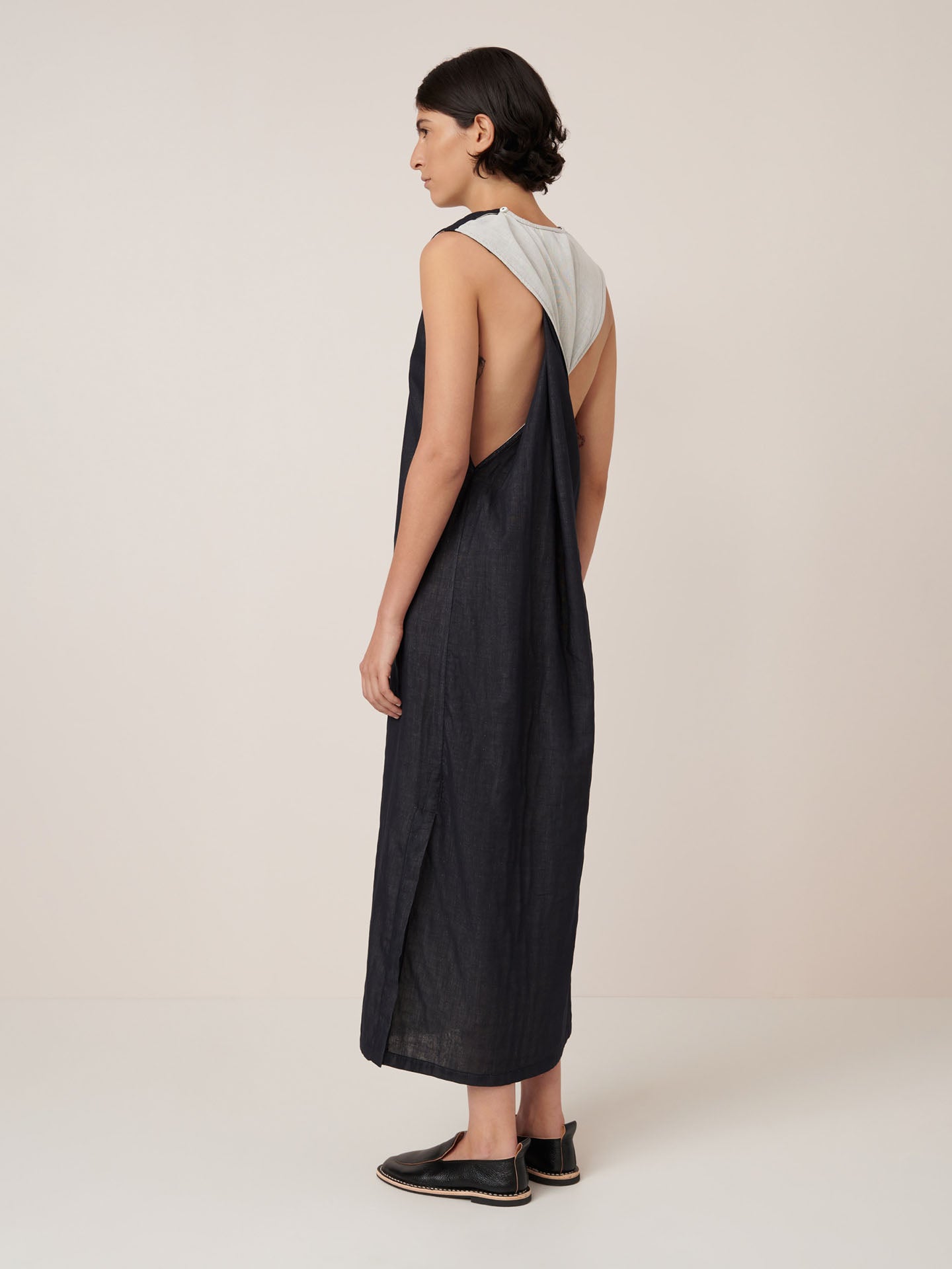 A person stands sideways wearing the Kowtow Ensemble Dress – Black Ecru, a standard fit, sleeveless, loose silhouette over a light gray top and black slip-on shoes. Please refer to the size guide for garment measurements.
