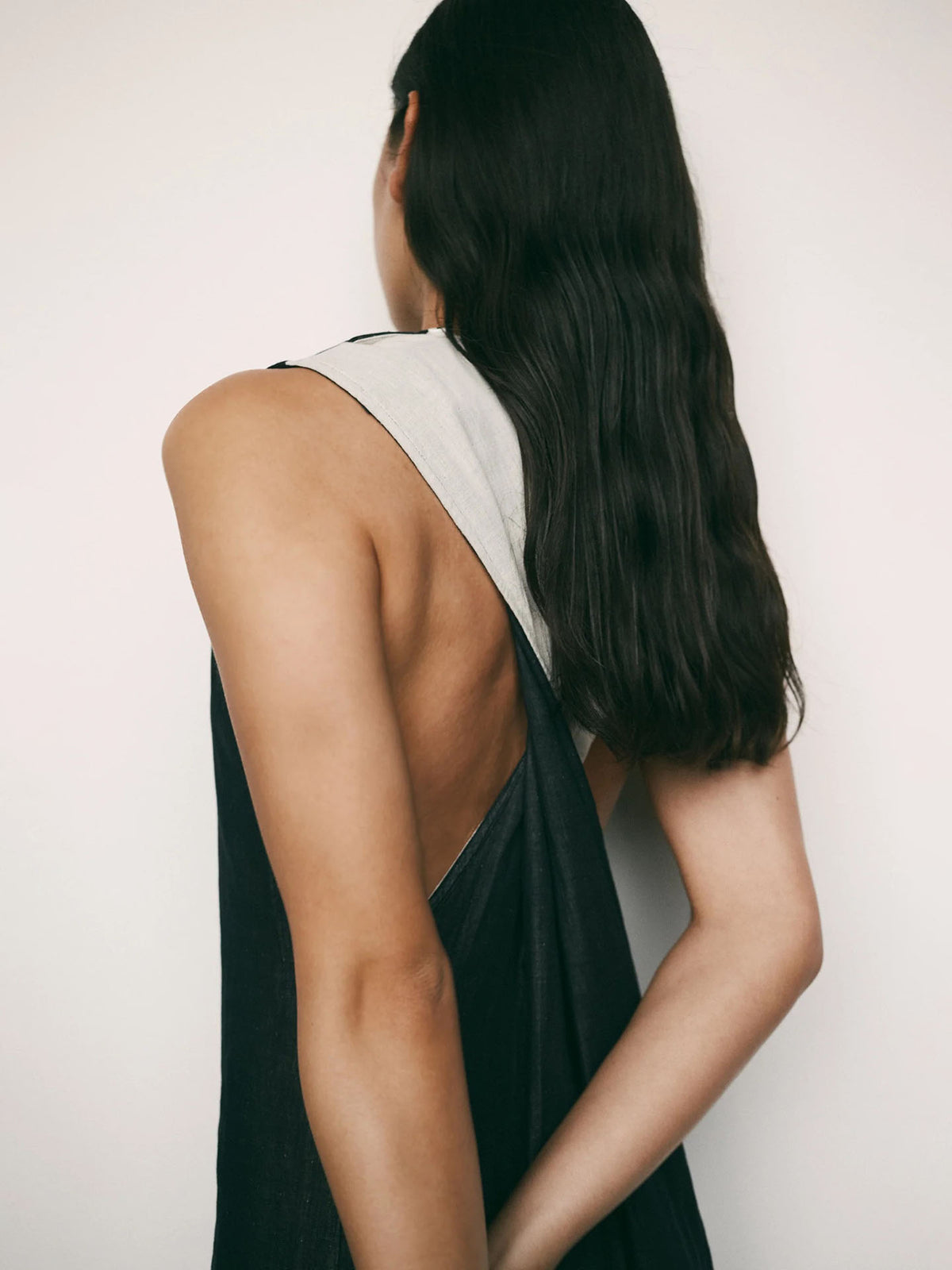 A person with long, dark hair faces away, wearing the Kowtow Ensemble Dress – Black Ecru in a standard fit, sleeveless style, with one arm bent behind their back against a plain background.