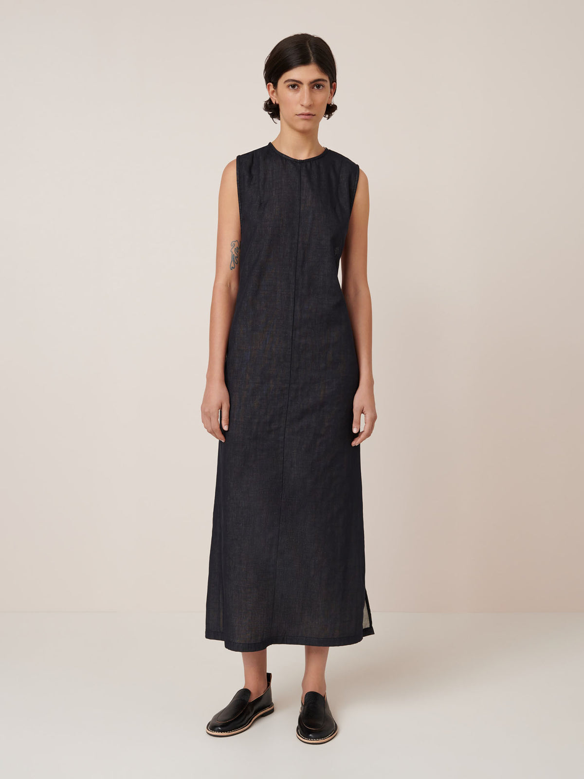 A person stands facing forward against a plain light background, wearing the Kowtow Ensemble Dress – Black Ecru, a standard fit sleeveless ankle-length black dress, and black slip-on shoes.