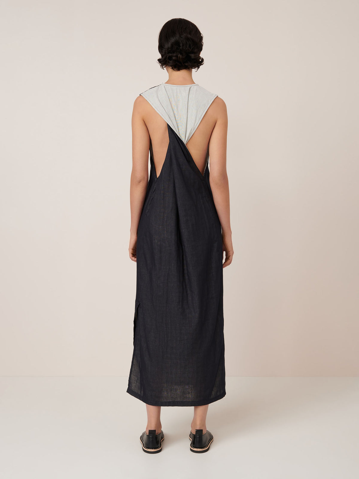 A person faces away, wearing Kowtow’s Ensemble Dress – Black Ecru, a sleeveless, backless style with a light gray upper panel, paired with slip-on shoes. The standard fit is shown against a plain, neutral background.