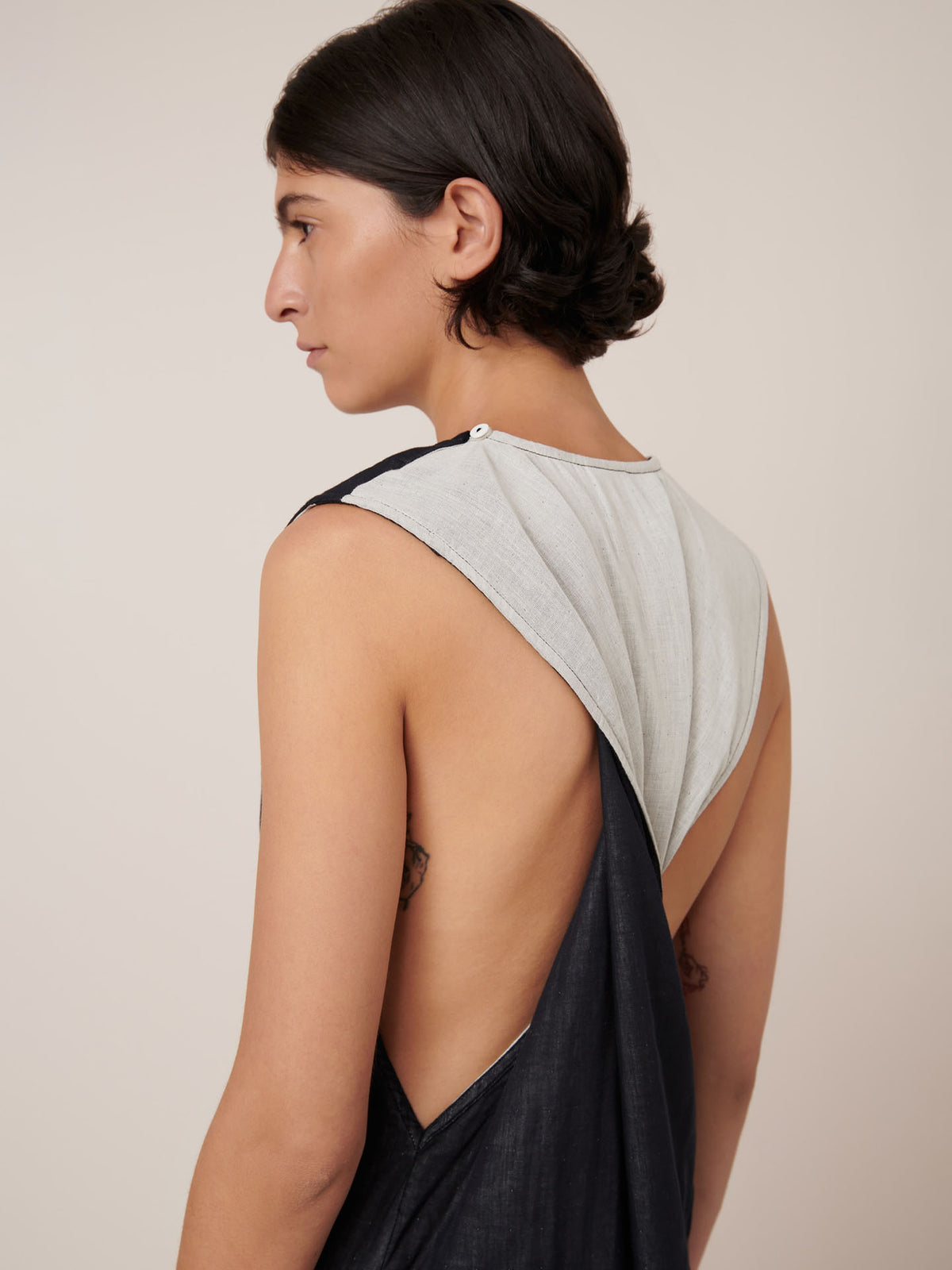 Model with short dark hair wears the Kowtow Ensemble Dress – Black Ecru, a sleeveless, backless, standard fit two-tone piece, standing sideways against a plain background.