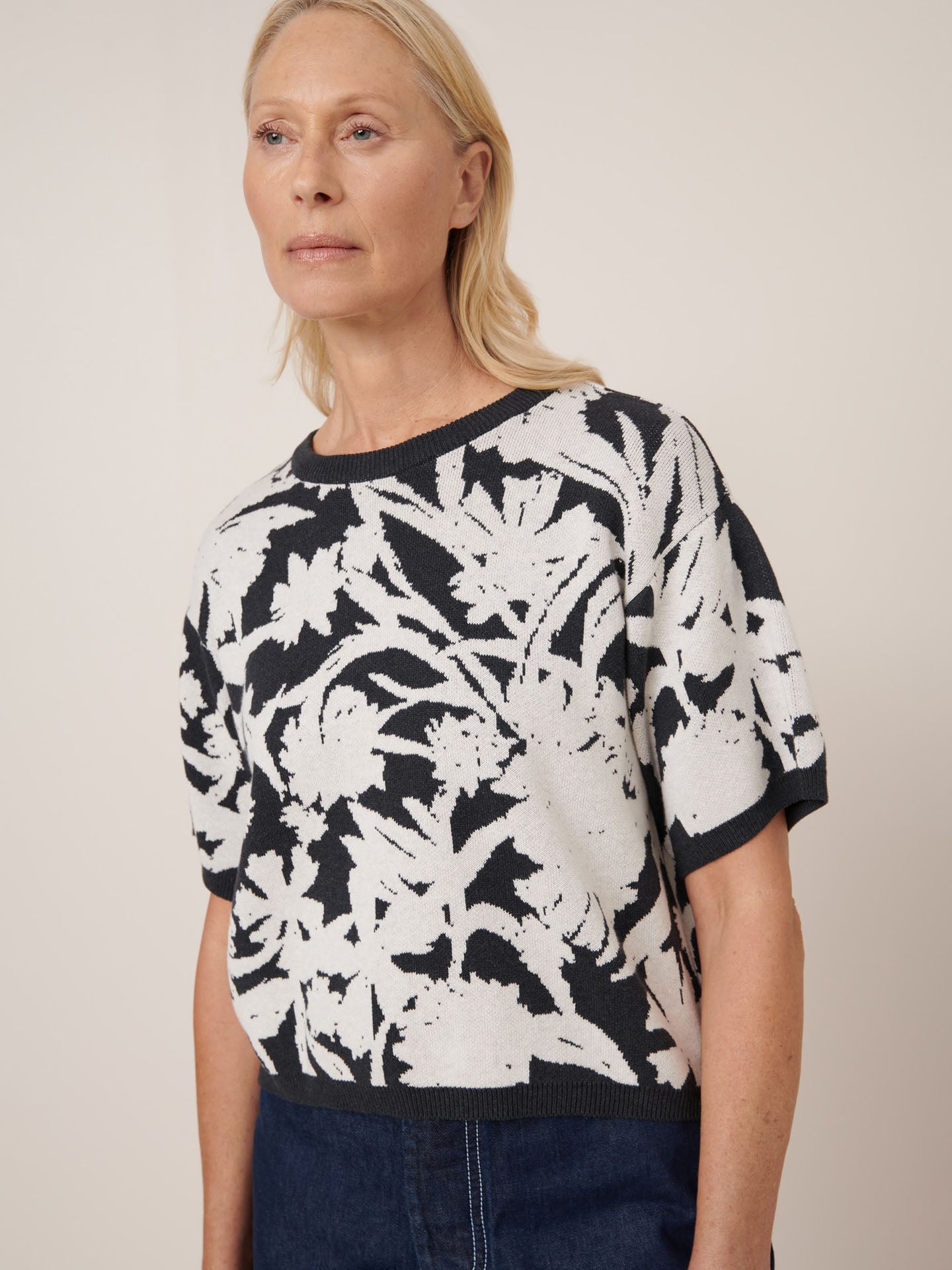 A woman wears the Kowtow Etched Top in Etched Jacquard—a relaxed-fit, short-sleeve black and white floral sweater—paired with dark blue jeans. Check our size guide for detailed garment measurements.