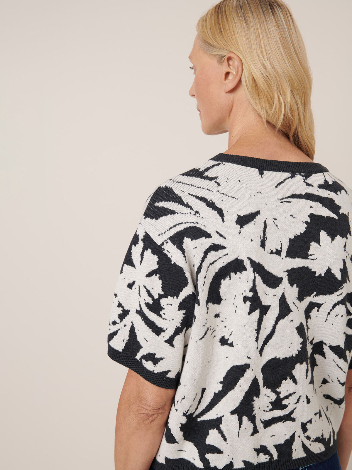 A person with blonde hair wears Kowtow&#39;s Etched Top in Etched Jacquard, featuring short sleeves and a bold black and white botanical pattern, shown from the back and side. Check the sizing guide for exact measurements.