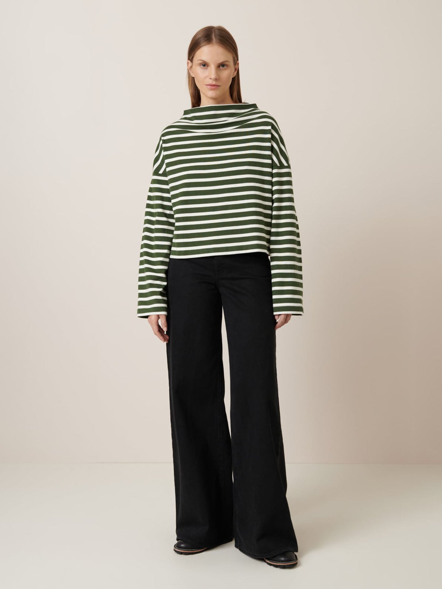 A woman faces forward in the Kowtow Field Top – Green Lily Stripe, a relaxed fit green and white striped long-sleeve, paired with black wide-leg pants against a plain background. Refer to the size guide for garment measurements.