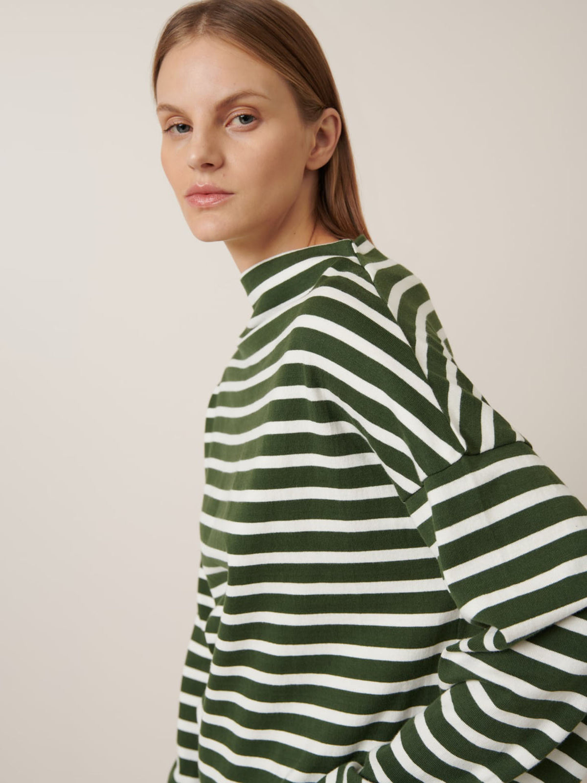 Model with straight, light brown hair wears the Kowtow Field Top – Green Lily Stripe, a relaxed-fit, green and white striped long-sleeve shirt, against a neutral background. See our size guide for detailed garment measurements.