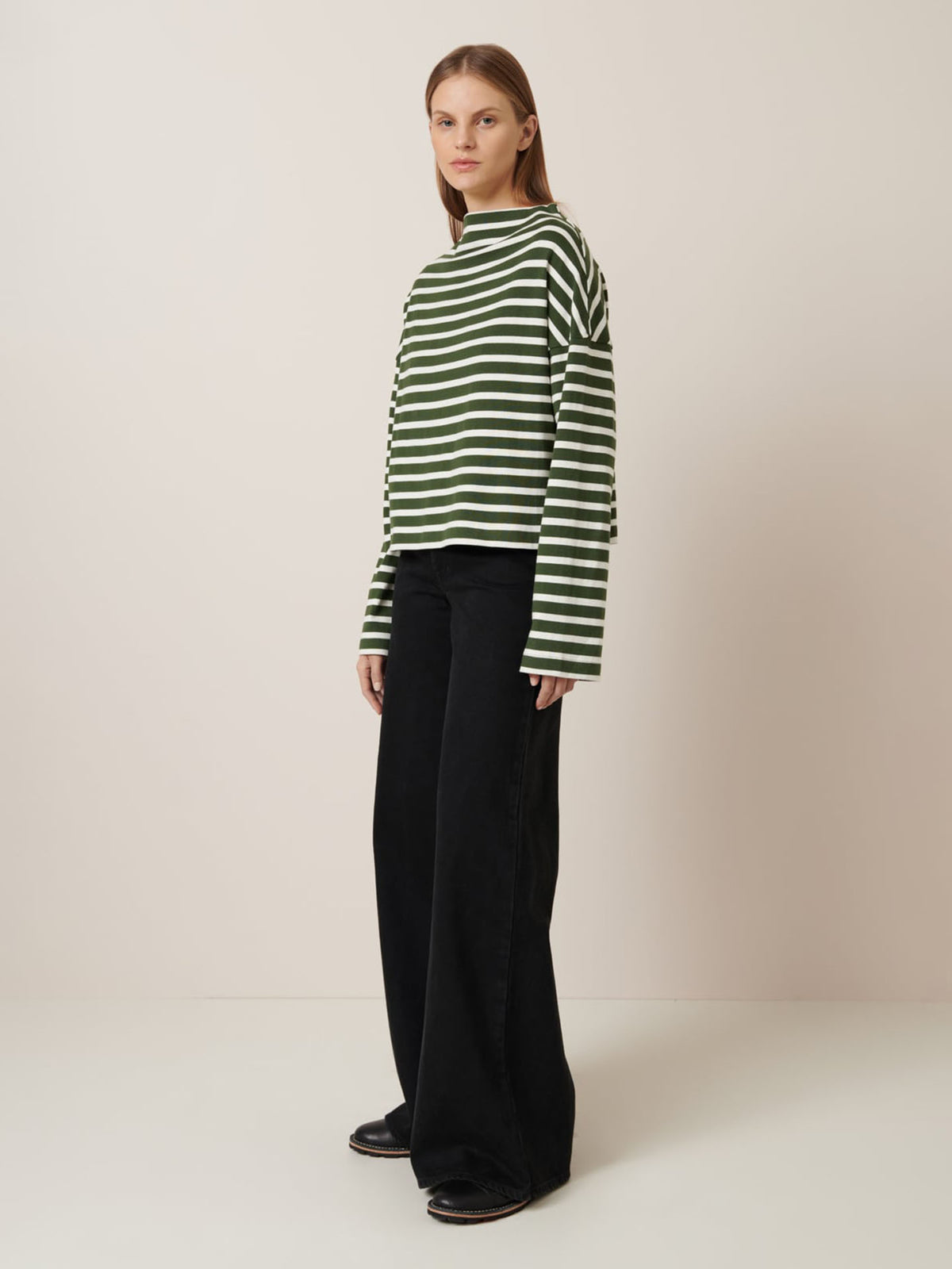 A woman models Kowtow&#39;s Field Top in Green Lily Stripe against a plain background, paired with loose black pants and black shoes to highlight the relaxed fit. For garment measurements, see our size guide.