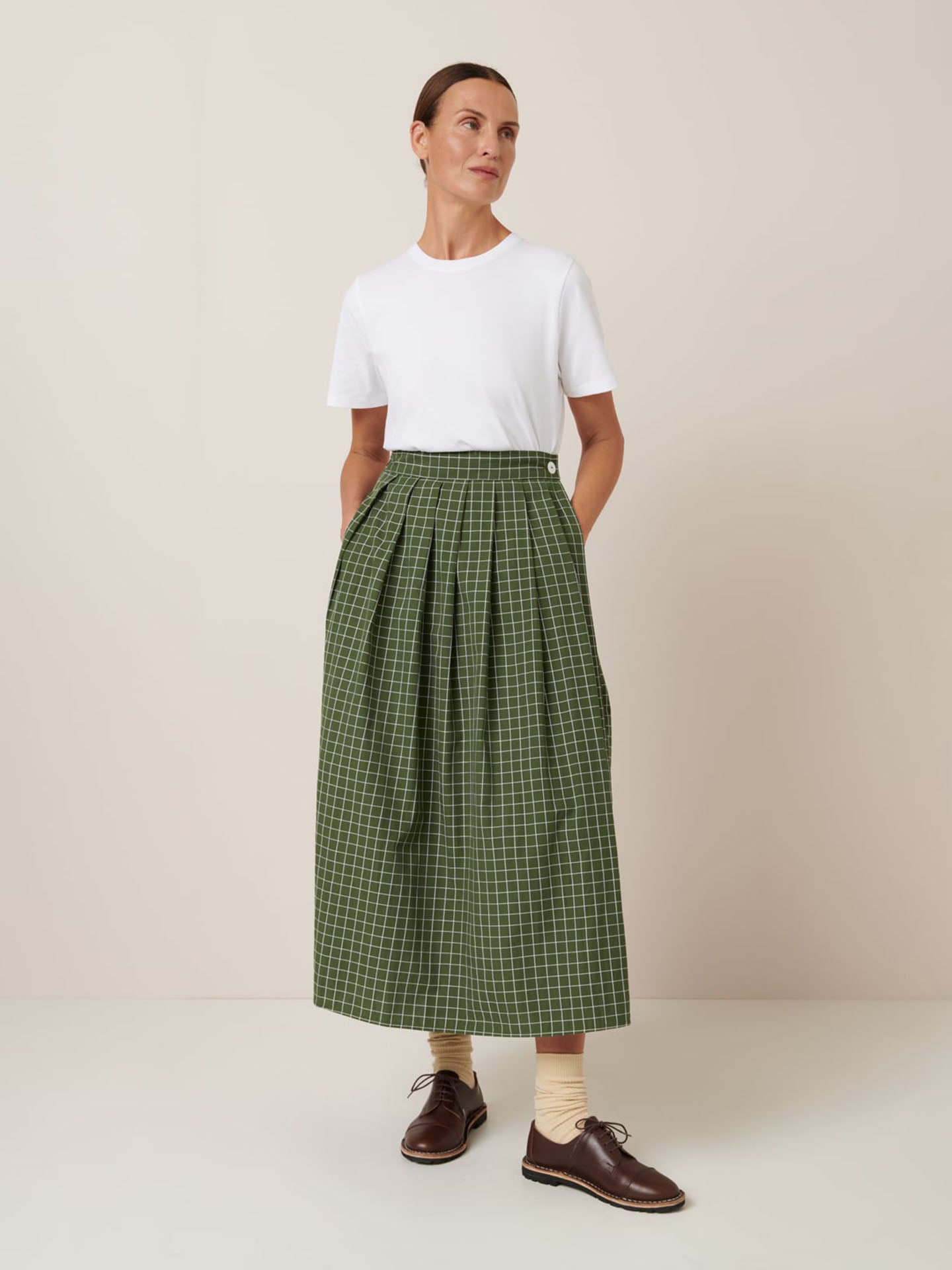 A person stands against a plain background wearing a white t-shirt, beige socks, brown shoes, and the Kowtow Flora Skirt – Forest Grid with a close fitting waistband—styled in their usual Kowtow size.