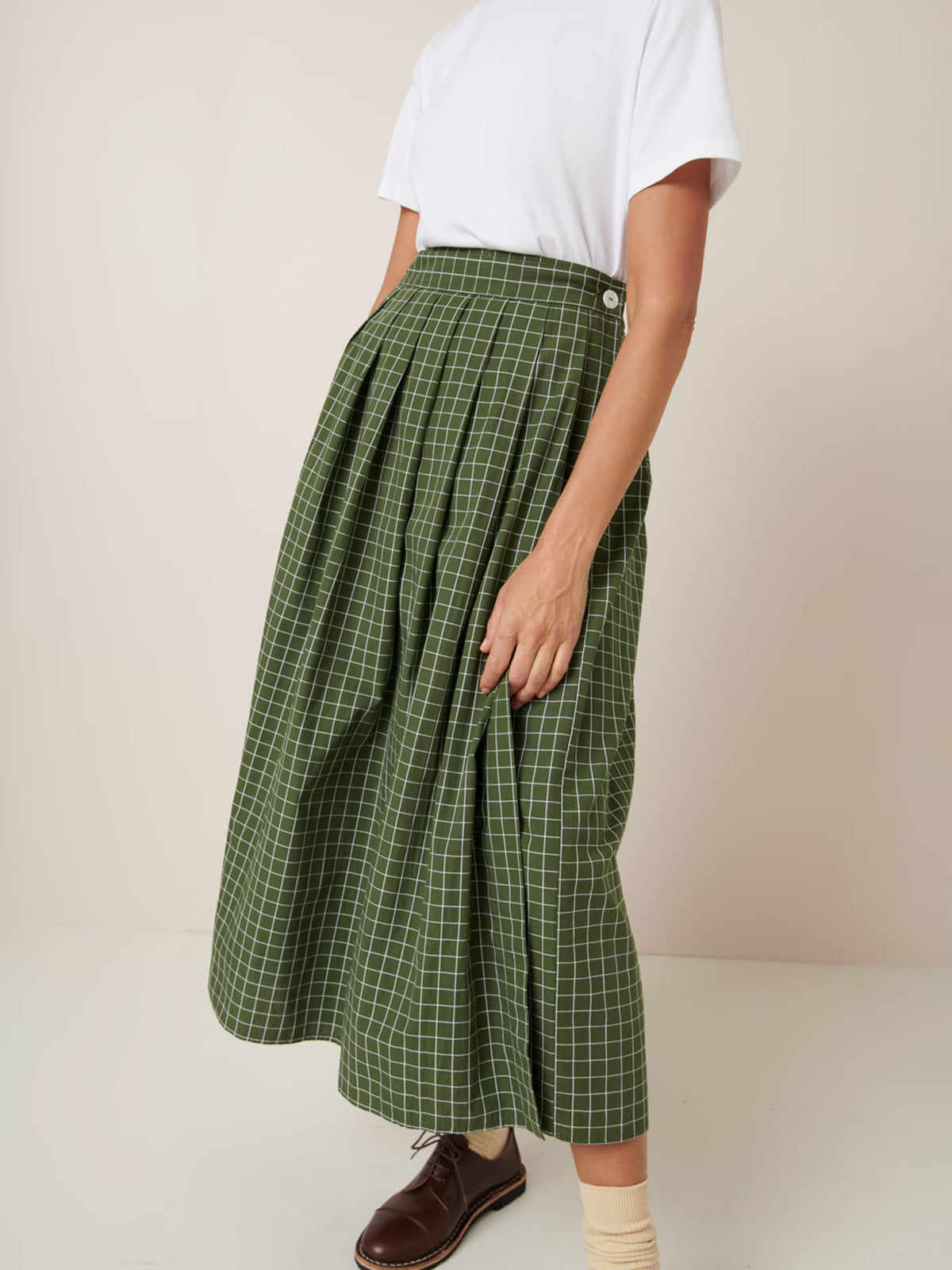 A person wears a white short-sleeve shirt, beige socks, brown shoes, and the Kowtow Flora Skirt – Forest Grid—a green checkered midi skirt with a close-fitting waistband—standing against a plain background.