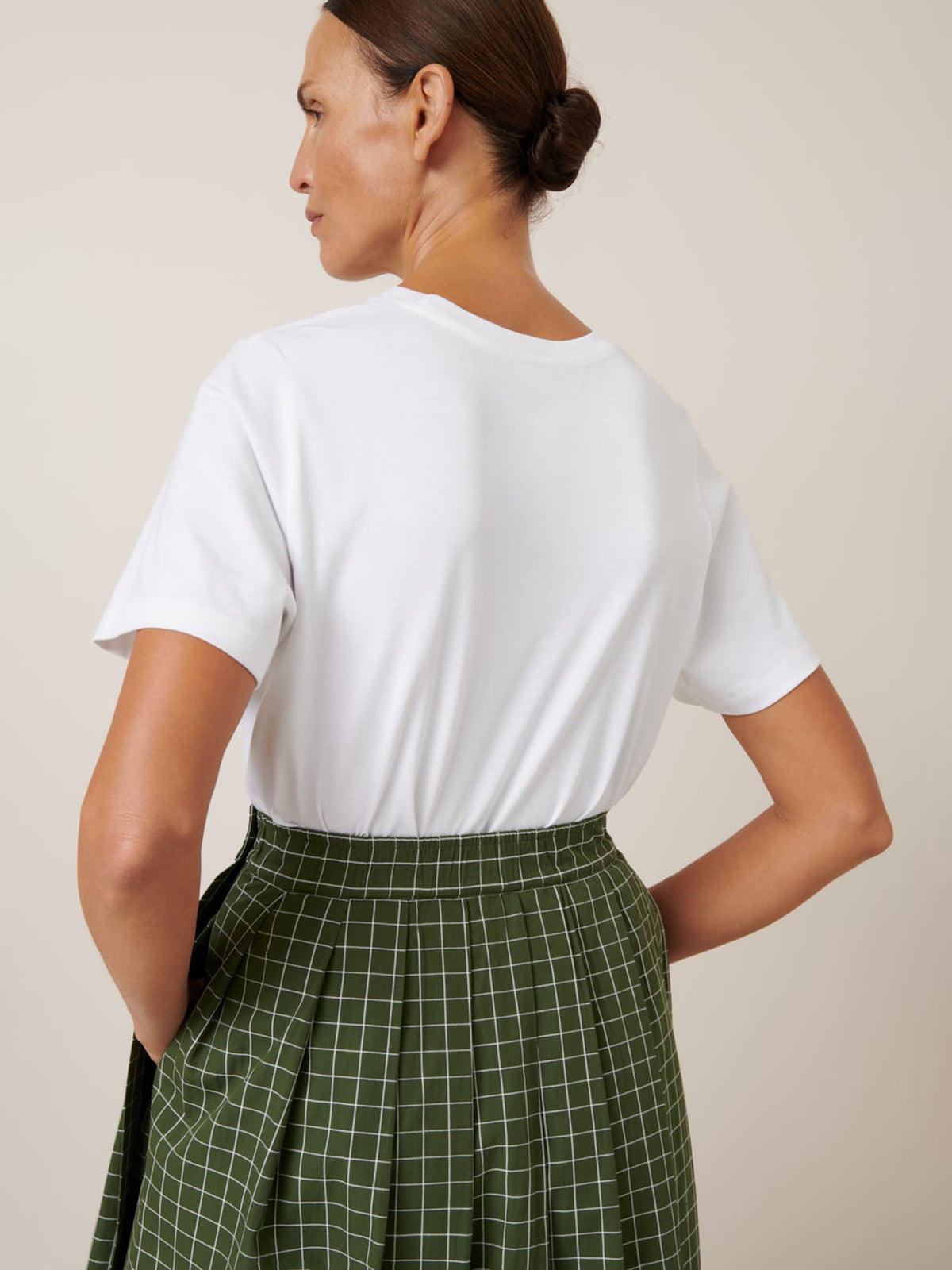 A woman faces away, wearing a white short-sleeve t-shirt tucked into the Kowtow Flora Skirt – Forest Grid, a high-waisted green skirt with a white grid pattern and close fitting waistband.