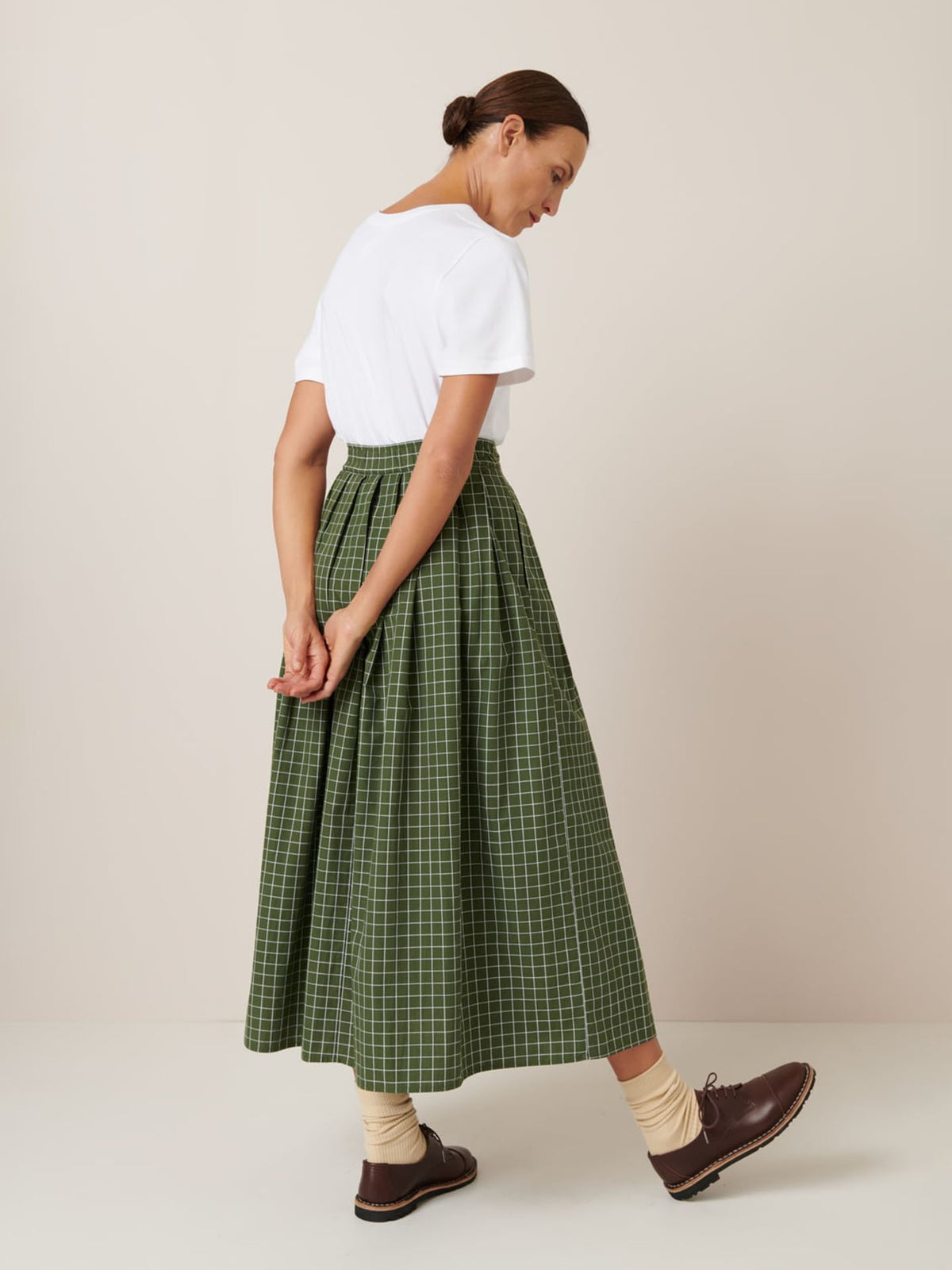 A person stands sideways against a plain background wearing a white t-shirt, beige socks, brown shoes, and the Kowtow Flora Skirt – Forest Grid, a green checkered midi skirt with a close-fitting waistband.