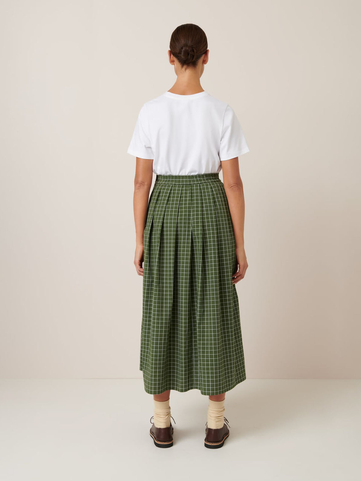A person stands facing away, wearing a standard fit white t-shirt, the Kowtow Flora Skirt – Forest Grid with a close fitting waistband, beige socks, and brown shoes against a plain background.