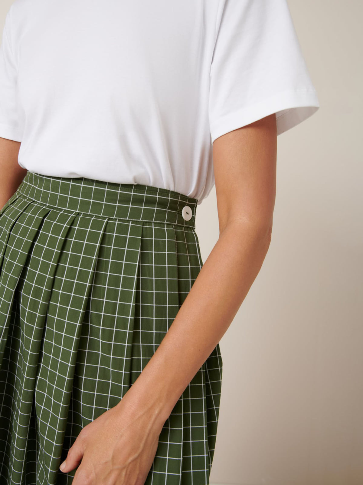 A person wears a white short-sleeve shirt and the Kowtow Flora Skirt – Forest Grid, a high-waisted green skirt with a white grid pattern, side button detail, close fitting waistband, and standard Kowtow fit.