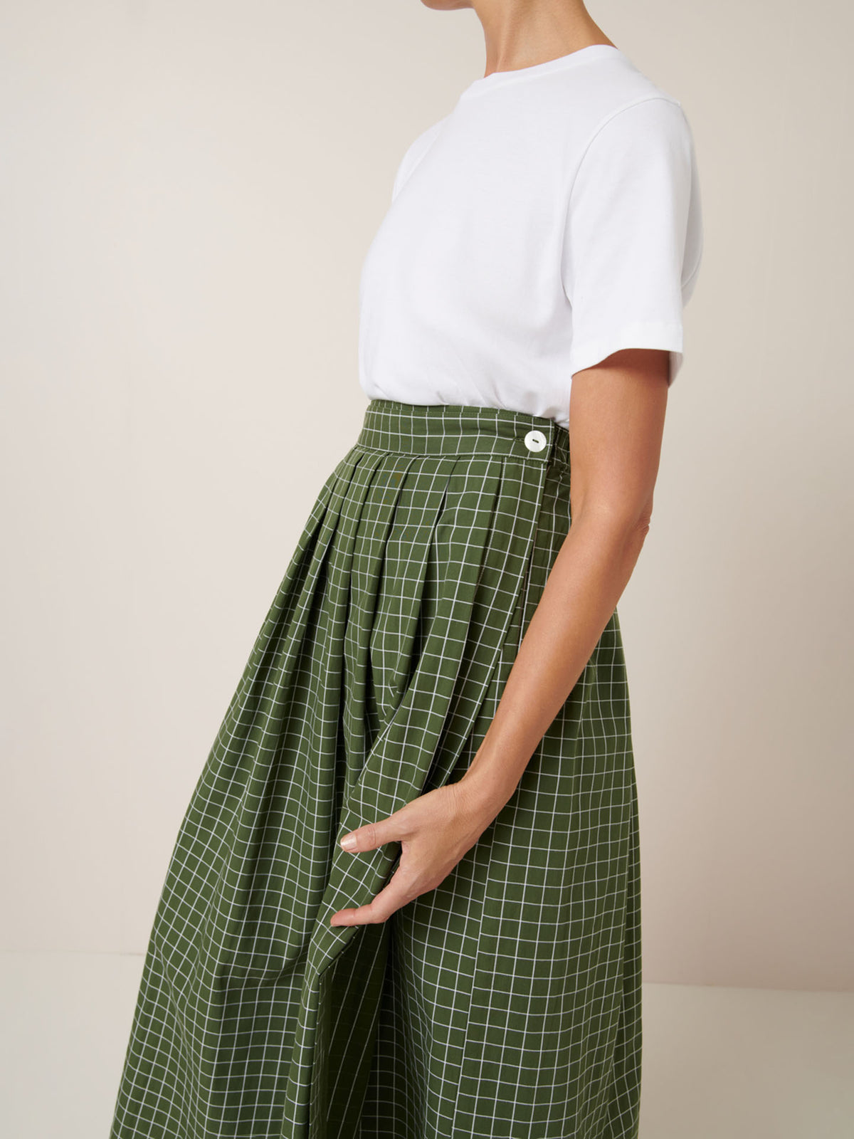 A person wears a plain white short-sleeve t-shirt tucked into the Kowtow Flora Skirt – Forest Grid, featuring a high-waisted fit, white grid pattern, button detail, and a close-fitting waistband for a standard look.