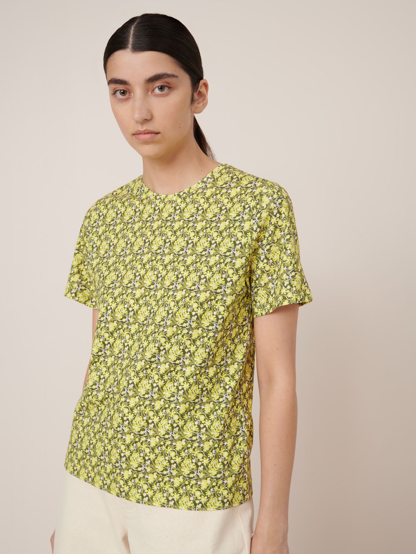 A person with long dark hair wears the Kowtow Flora Tee – Flora, a yellow and green floral-patterned short-sleeve shirt paired with light pants, standing against a plain beige background to showcase standard fit for easy garment measurements.