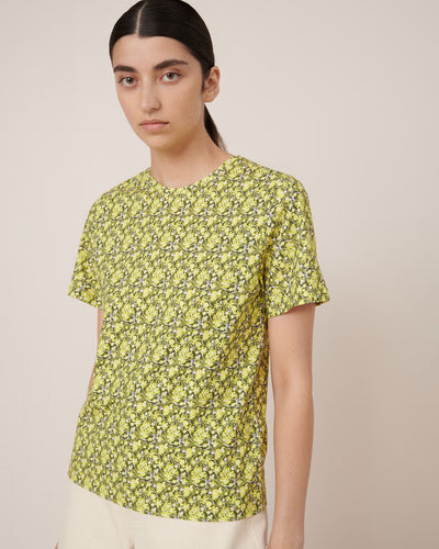 A person with long dark hair wears the Kowtow Flora Tee – Flora, a yellow and green floral-patterned short-sleeve shirt paired with light pants, standing against a plain beige background to showcase standard fit for easy garment measurements.