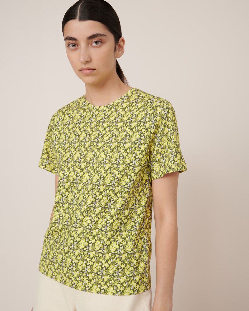A person with long dark hair wears the Kowtow Flora Tee – Flora, a yellow and green floral-patterned short-sleeve shirt paired with light pants, standing against a plain beige background to showcase standard fit for easy garment measurements.