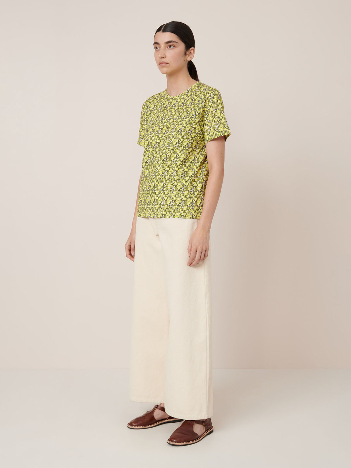 A person stands against a plain background wearing the Kowtow Flora Tee – Flora, paired with wide-leg cream pants and brown flats—a simple example of standard fit. Check our size guide for exact Flora Tee garment measurements.