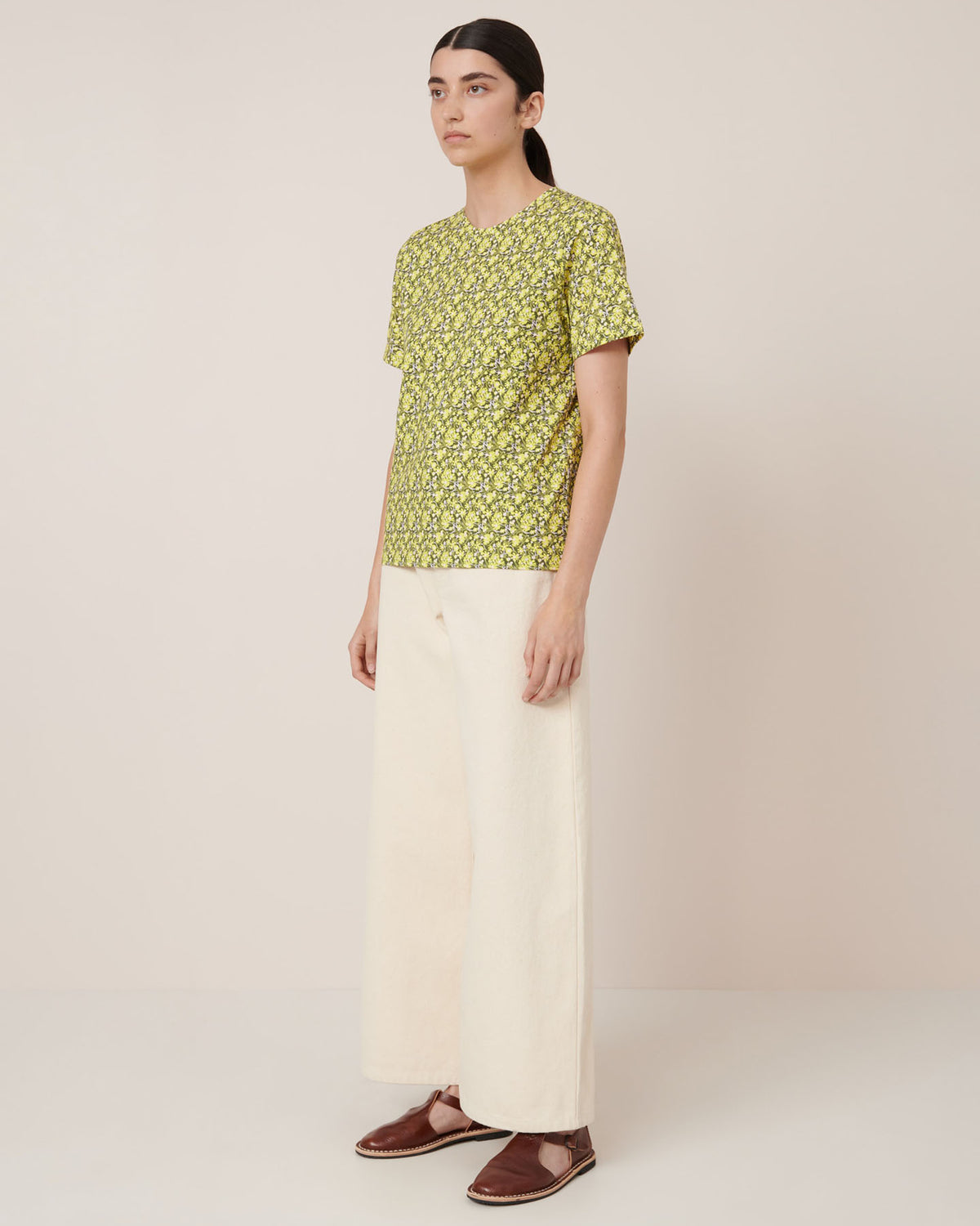 A person stands against a plain background wearing the Kowtow Flora Tee – Flora, paired with wide-leg cream pants and brown flats—a simple example of standard fit. Check our size guide for exact Flora Tee garment measurements.