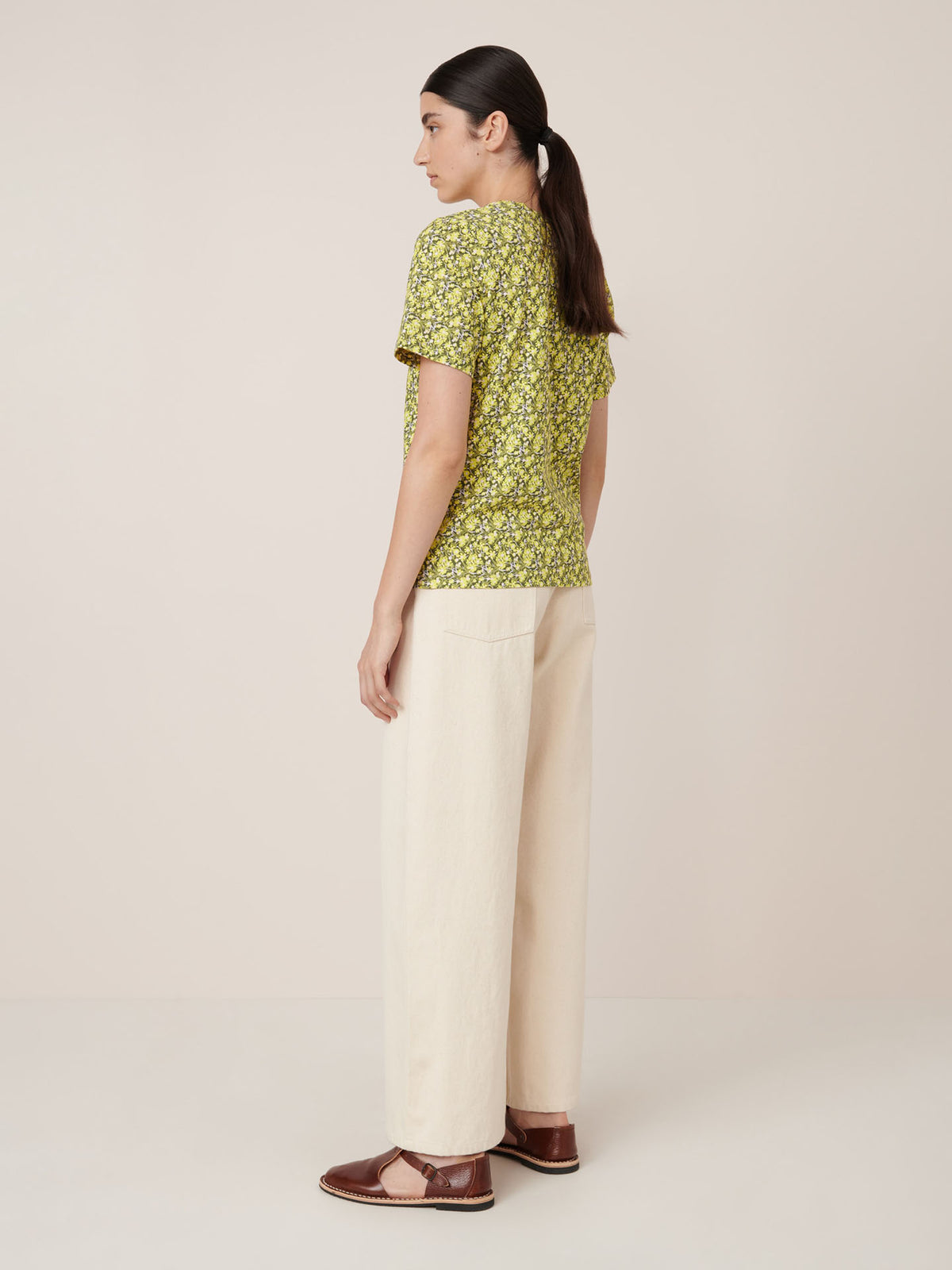 A woman stands sideways in the Kowtow Flora Tee – Flora, cream wide-leg pants, and brown flats against a plain background, showcasing a standard fit. Check our size guide for precise measurements.