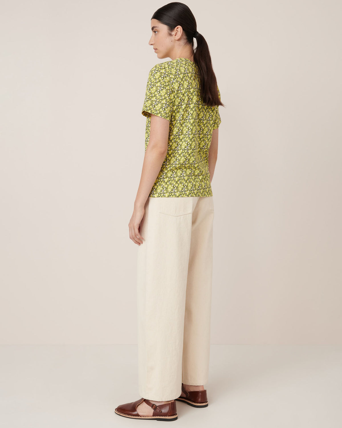 A woman stands sideways in the Kowtow Flora Tee – Flora, cream wide-leg pants, and brown flats against a plain background, showcasing a standard fit. Check our size guide for precise measurements.