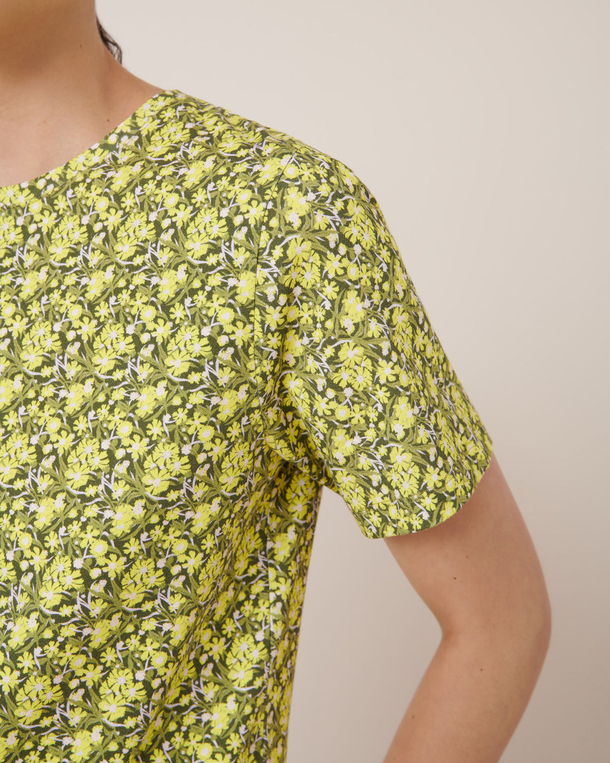 Close-up of a person wearing the Kowtow Flora Tee in a yellow and green floral print, standing against a plain light background. See our size guide for garment measurements and fit details.