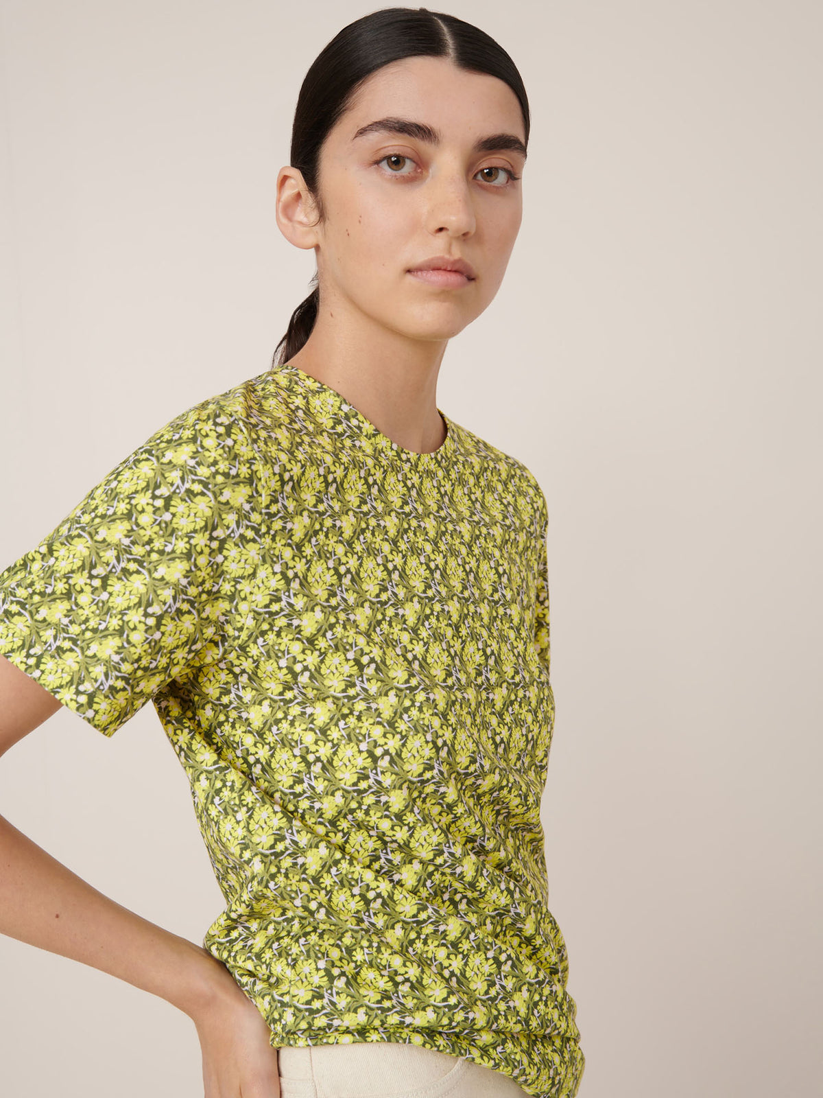 Person with long dark hair wears the Kowtow Flora Tee – Flora, a yellow floral short-sleeve shirt, paired with light pants against a plain background—ideal for displaying the fit and details of this standout top.